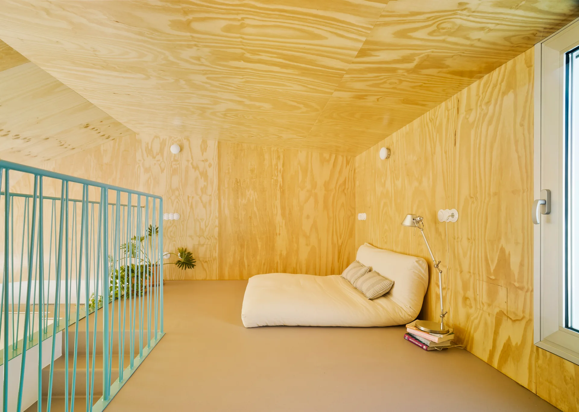 A low-profile floor mattress sits in an attic bedroom lined with natural plywood walls and ceilings next to a teal-colored metal staircase railing.