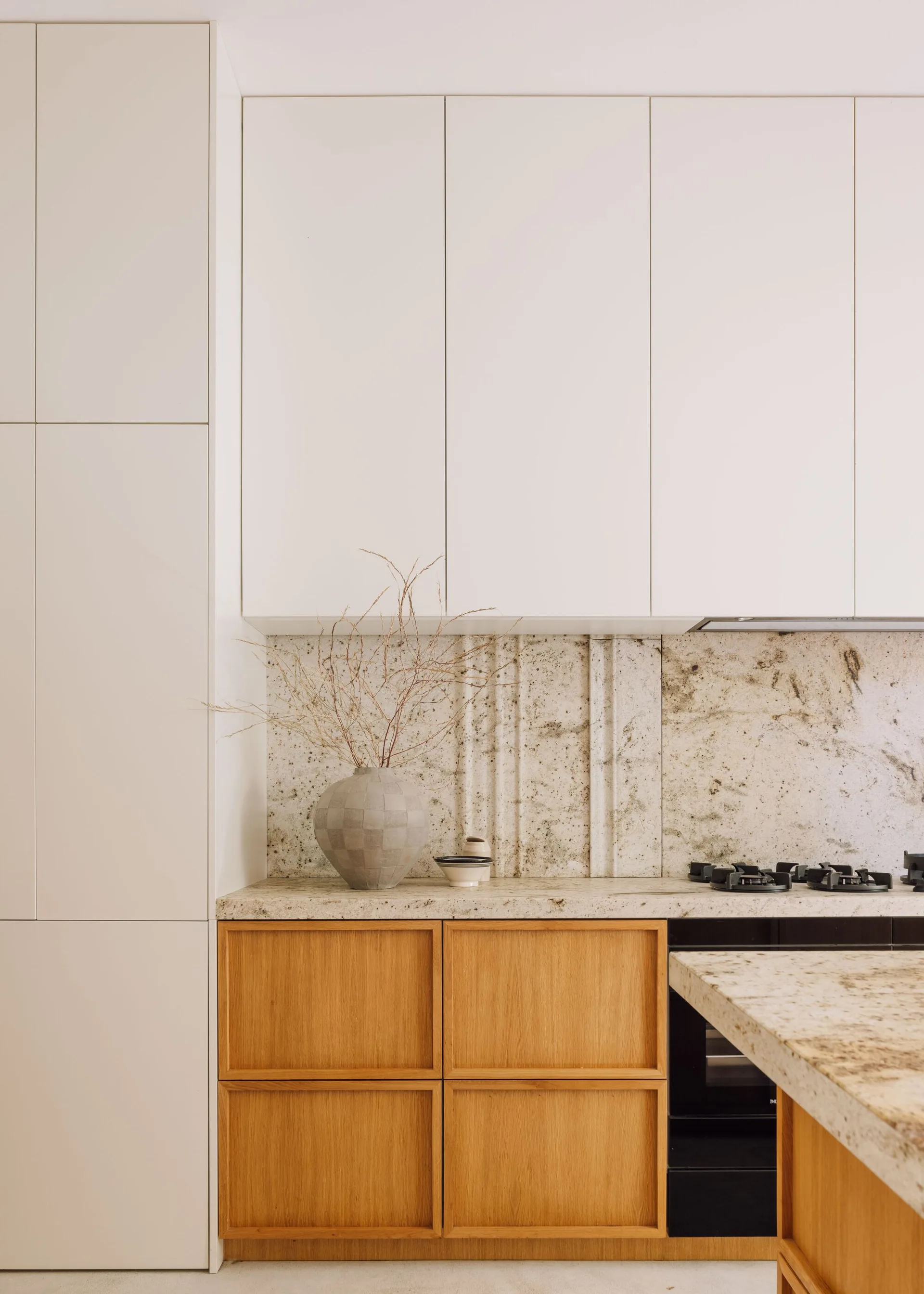 A minimalist kitchen features off-white upper cabinets, natural wood lower cabinets, and a textured stone countertop and backsplash.