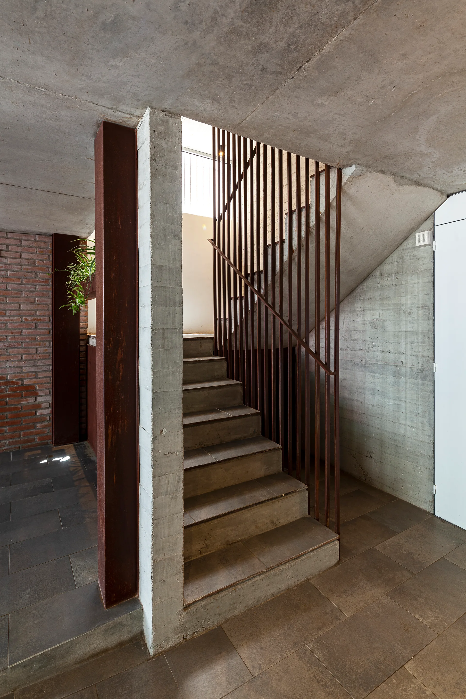 An industrial interior staircase constructed from raw concrete steps, featuring vertical rusted metal rail bars, concrete walls, and a brick wall.