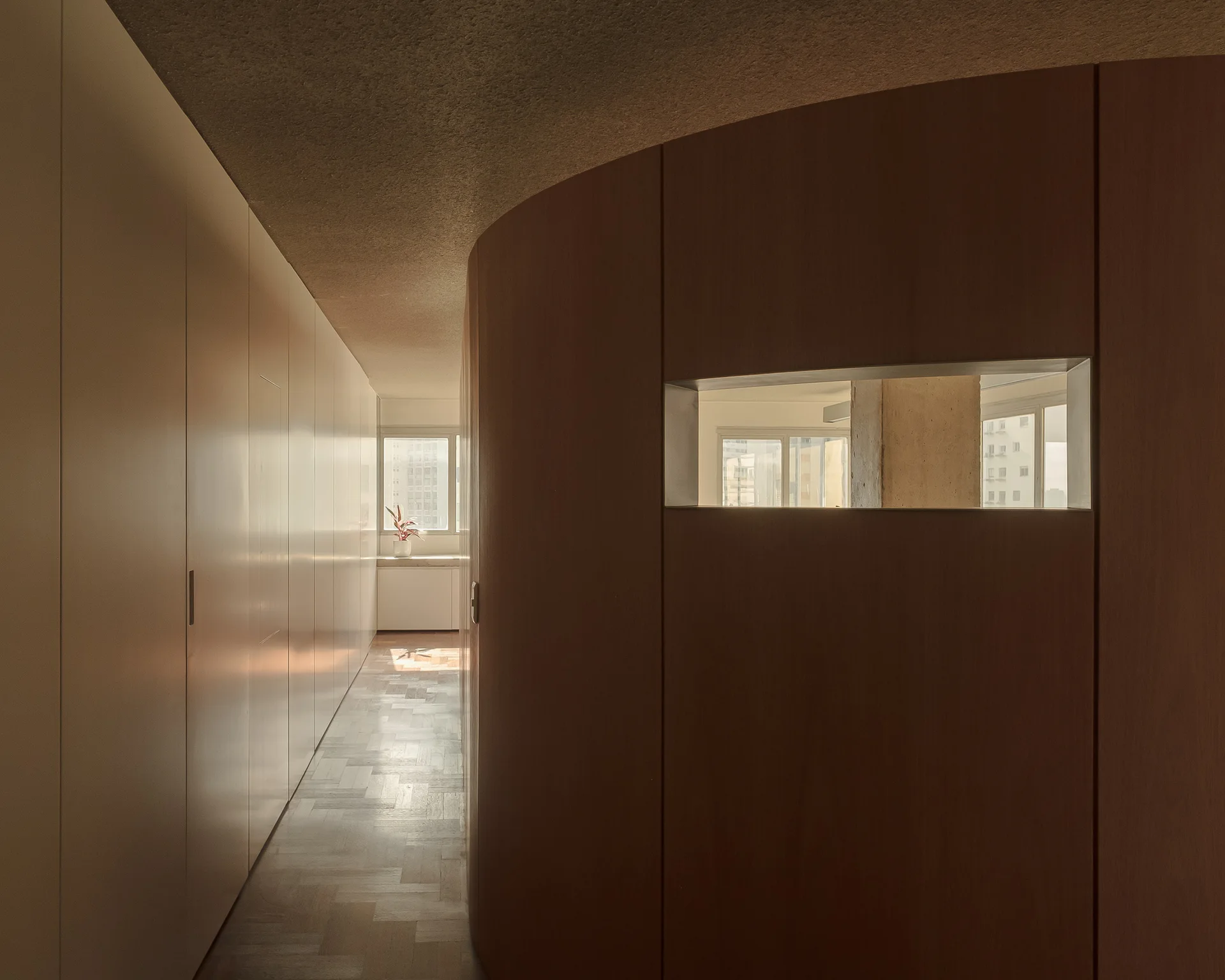 A minimalist hallway features a curved wood-paneled wall with a window cutout, herringbone parquet flooring, and a long run of seamless built-in cabinets.