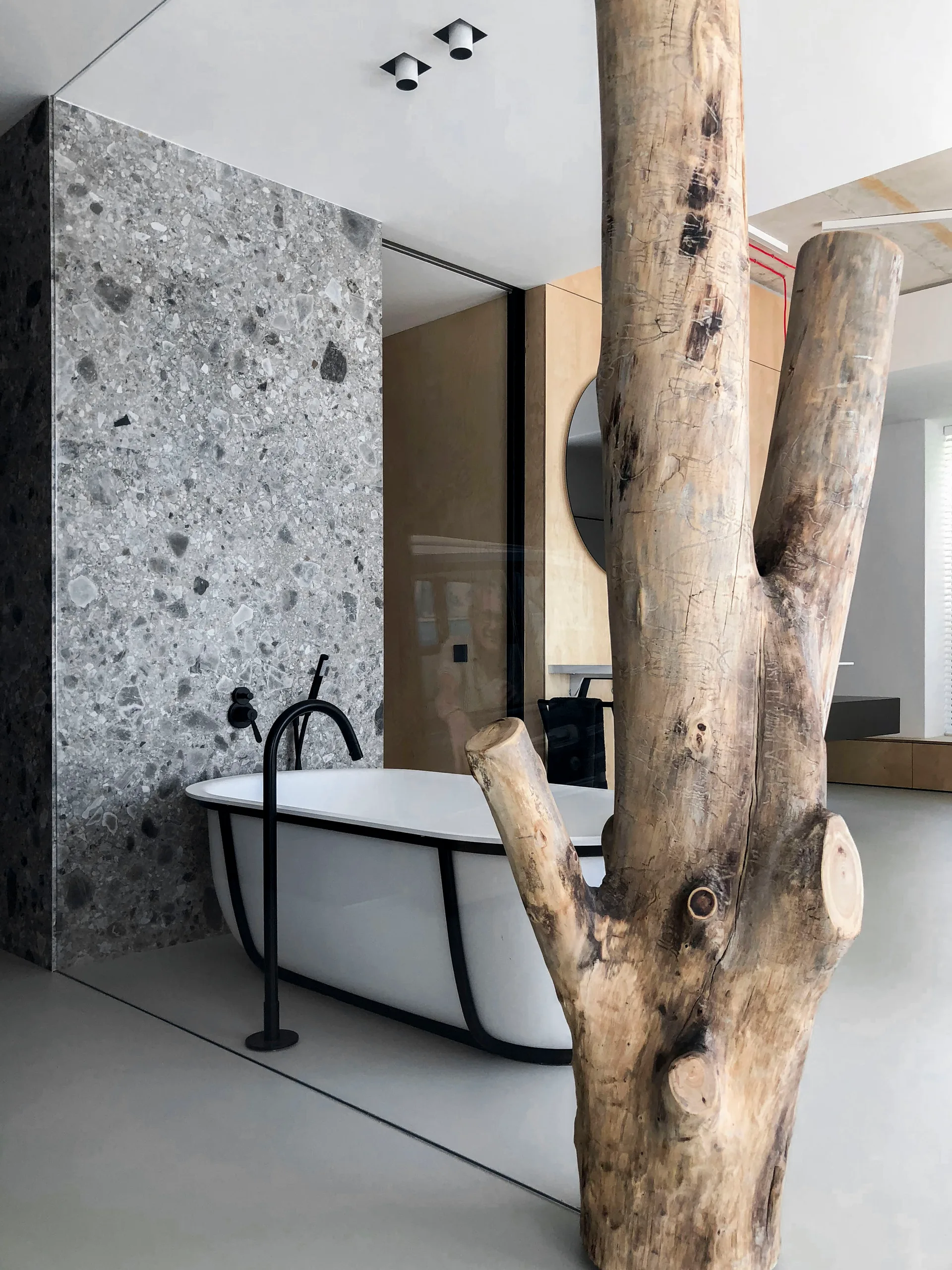 A modern bathroom features a white freestanding bathtub with a black frame against a gray terrazzo wall, partially framed by a raw tree trunk column.