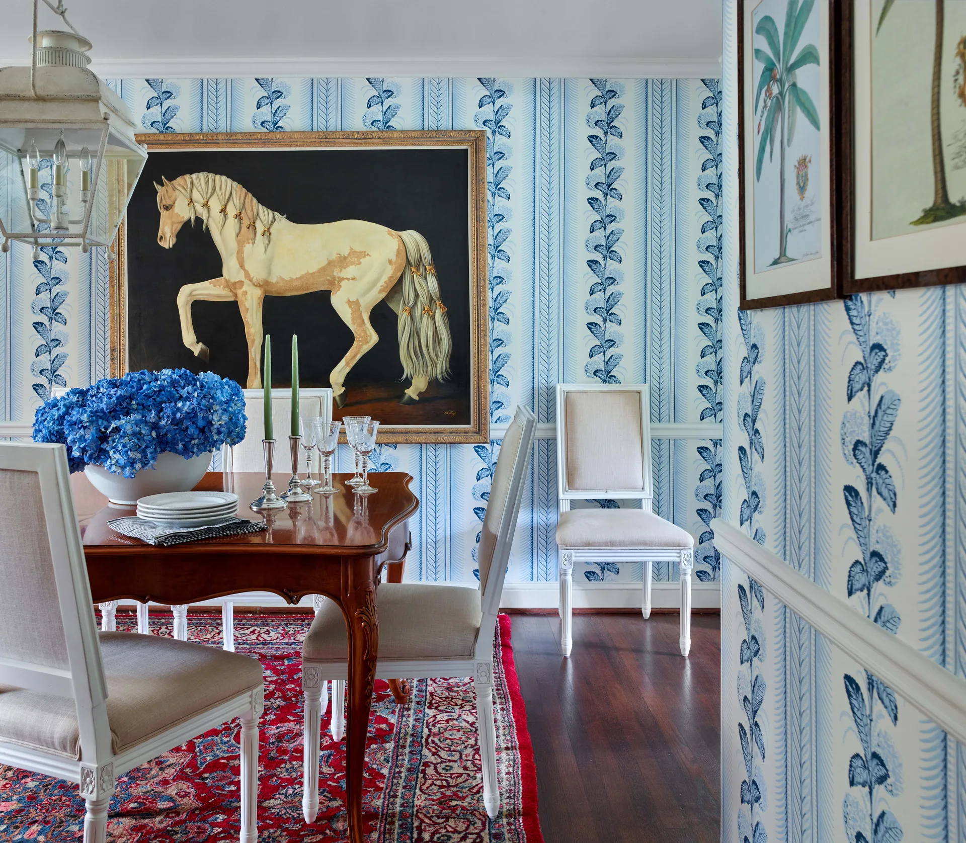 A traditional dining room with a dark wood table, white upholstered chairs, blue and white striped wallpaper, and a large framed horse painting.