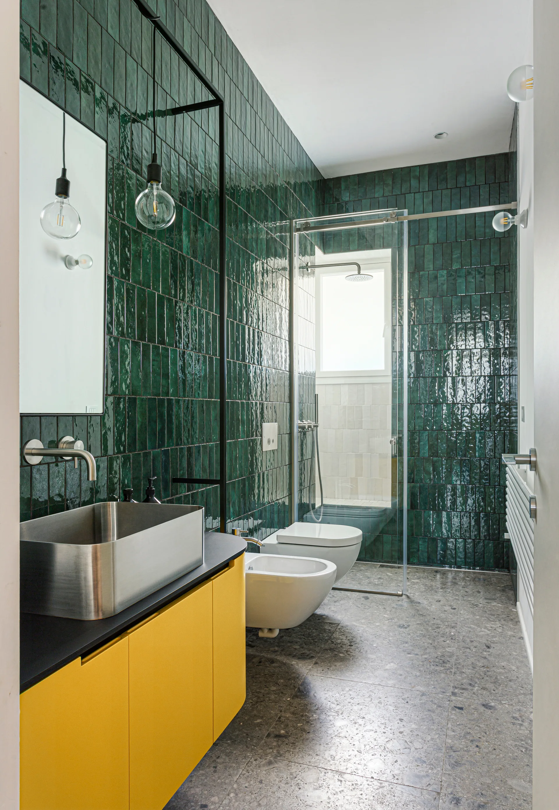 A modern bathroom features glossy green vertical tiles, a matte yellow vanity with a stainless steel sink, and gray terrazzo flooring.