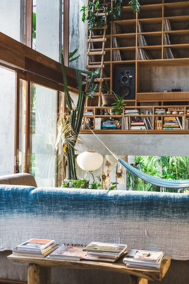 A living room features a blue ombre textured sofa, a low wood bench, and a tall floor-to-ceiling wooden bookshelf set against a concrete wall.