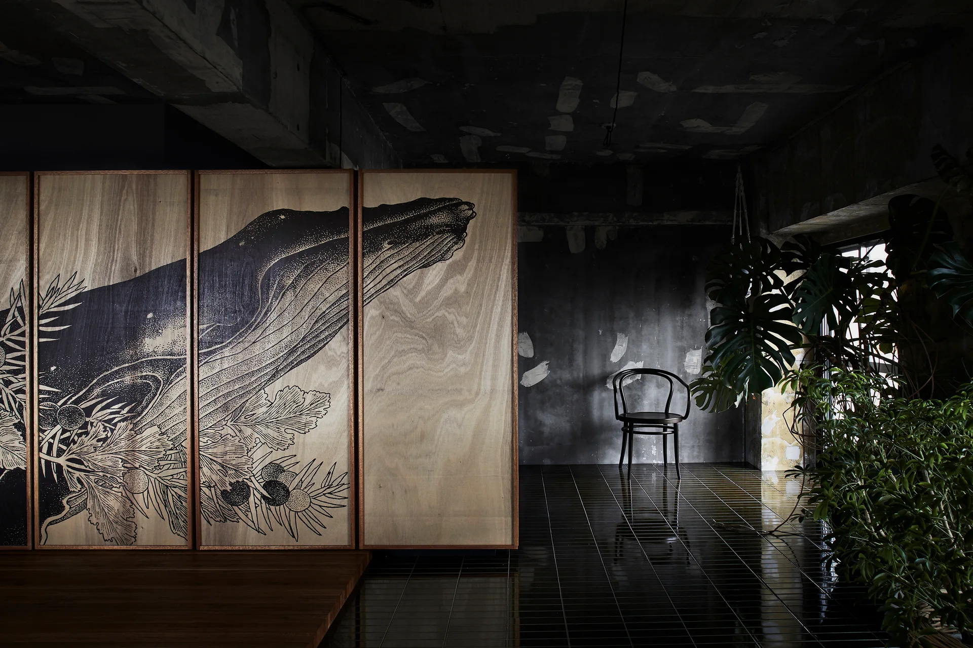 A wooden room divider with a graphic whale print sits on a glossy dark tiled floor beside raw concrete walls and green houseplants.