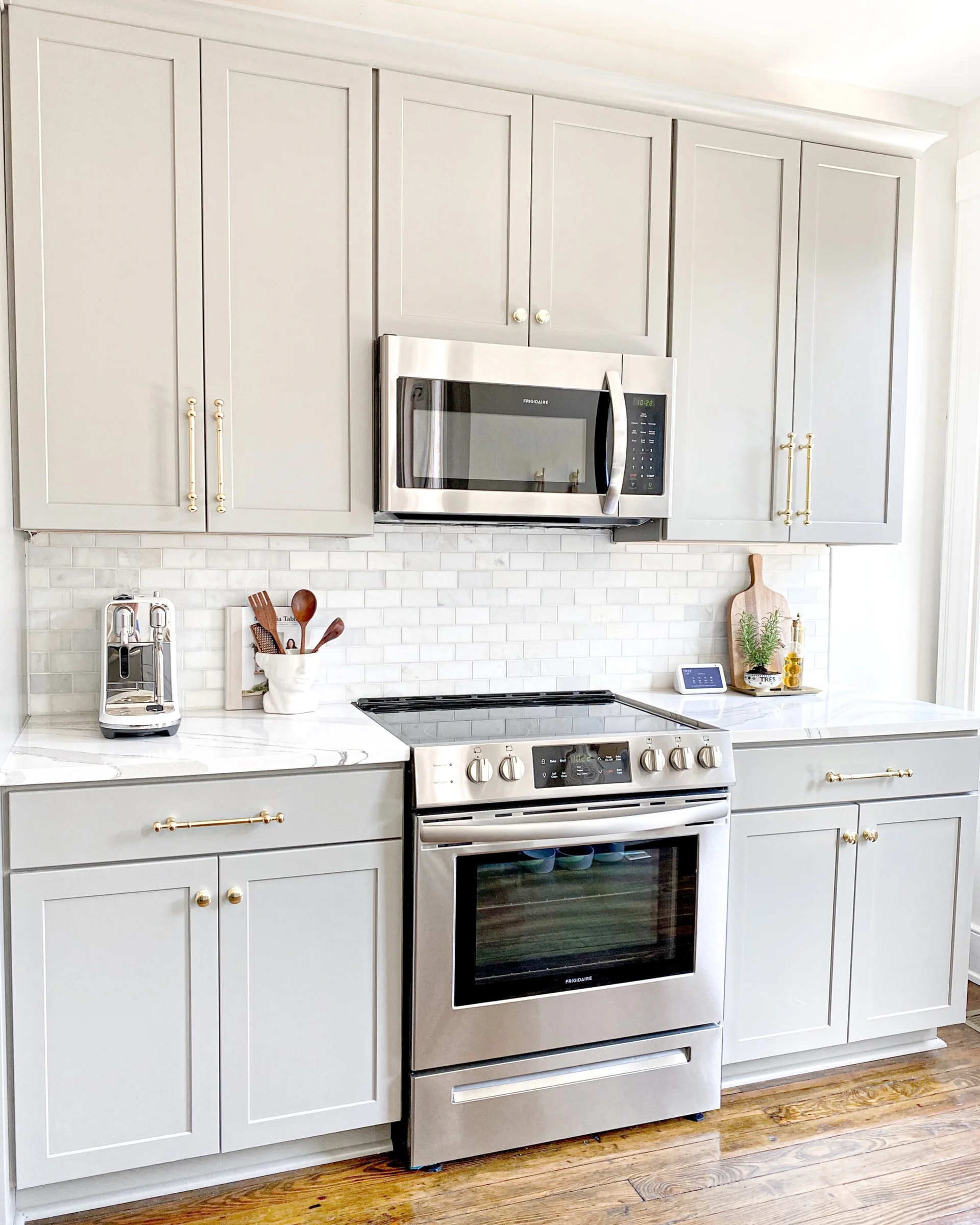 A bright kitchen features light gray shaker cabinets with gold handles, a white marble-look countertop, and stainless steel appliances.