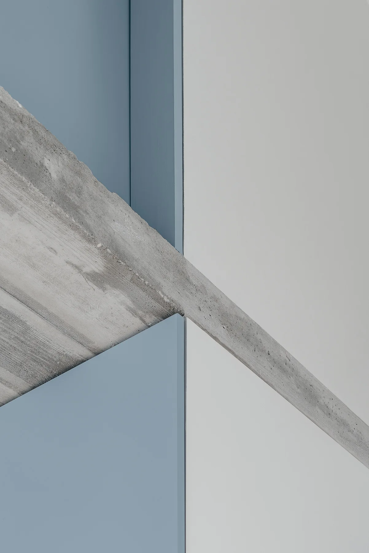An abstract architectural detail of a rough grey concrete beam intersecting with smooth, matte-finished dusty blue and off-white painted wall panels.