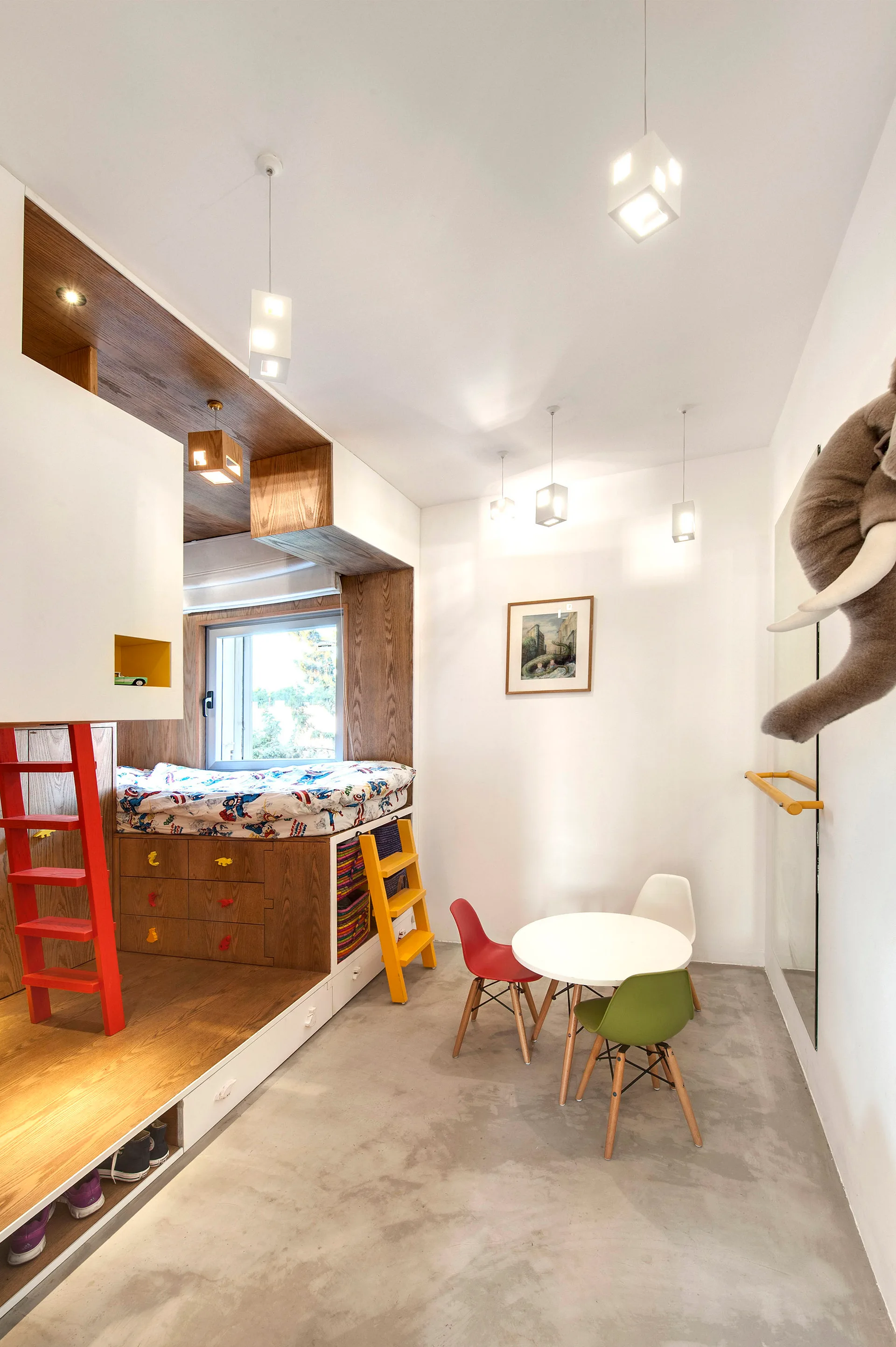 A children's bedroom features a built-in wooden platform bed with red and yellow ladders, a small round play table with colorful chairs, and a concrete floor.