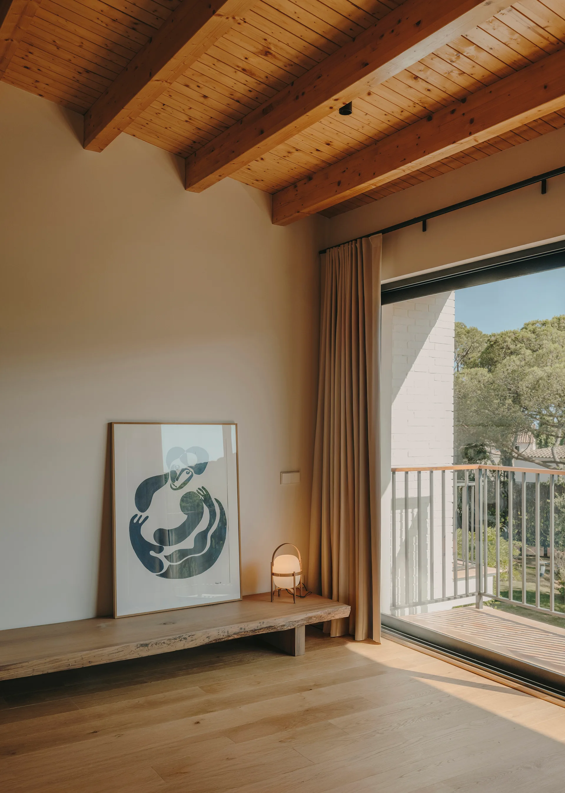 A sunlit minimalist room features an exposed wood beam ceiling, a low live-edge wooden bench with framed art, and a large sliding glass door.