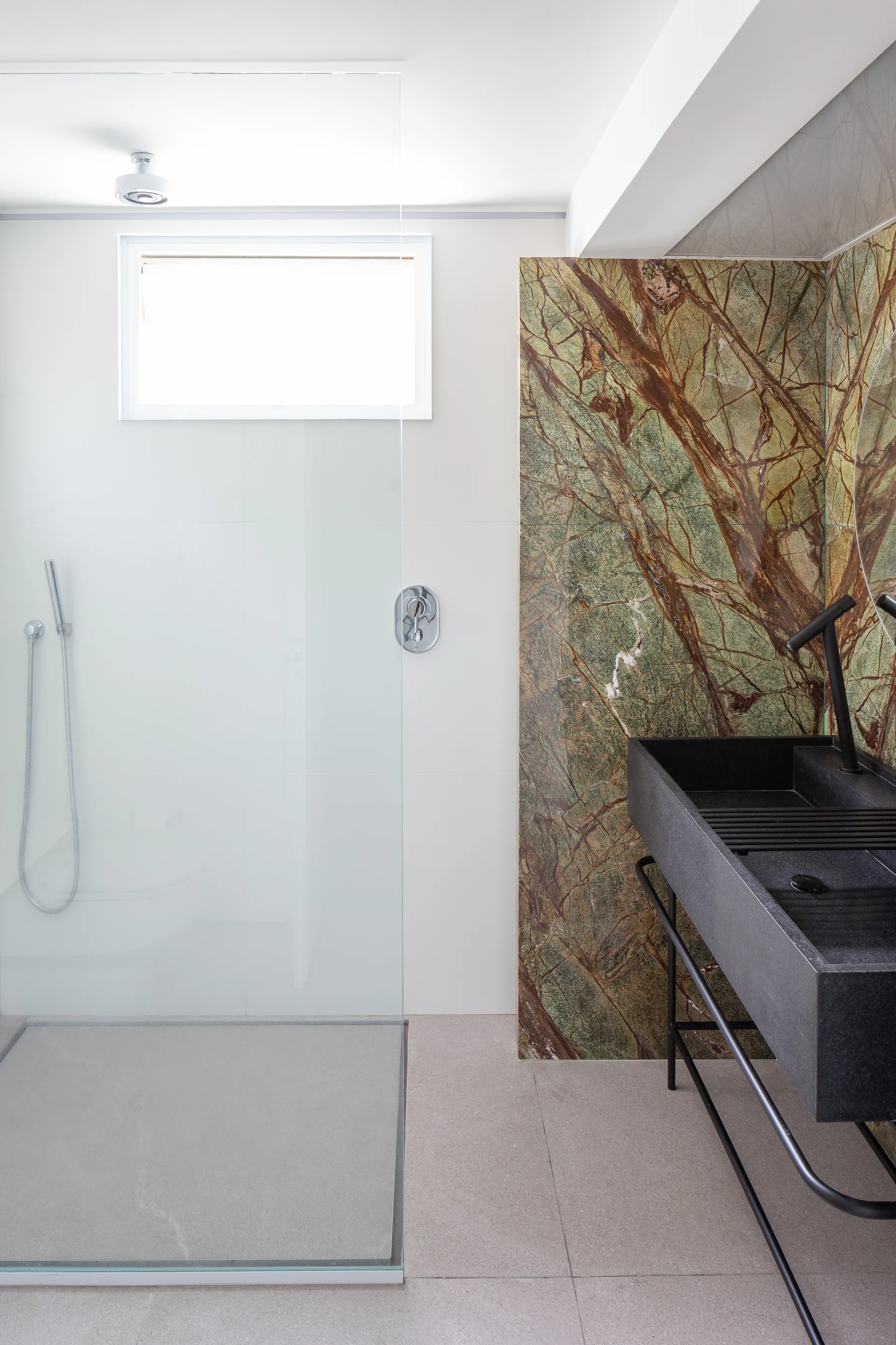 A modern bathroom features a black rectangular sink on a metal frame set against a green veined stone accent wall next to a glass-enclosed shower.