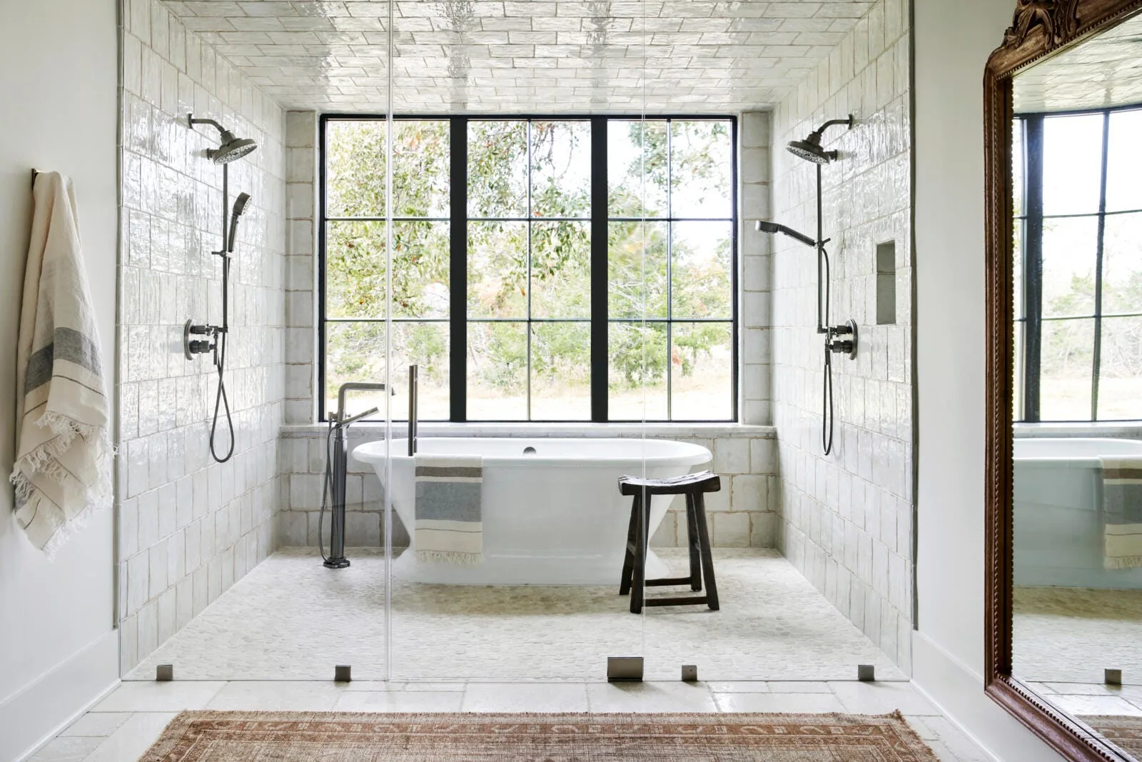 A white-tiled wet room features a freestanding oval bathtub centered in front of a black grid window, flanked by two shower heads.