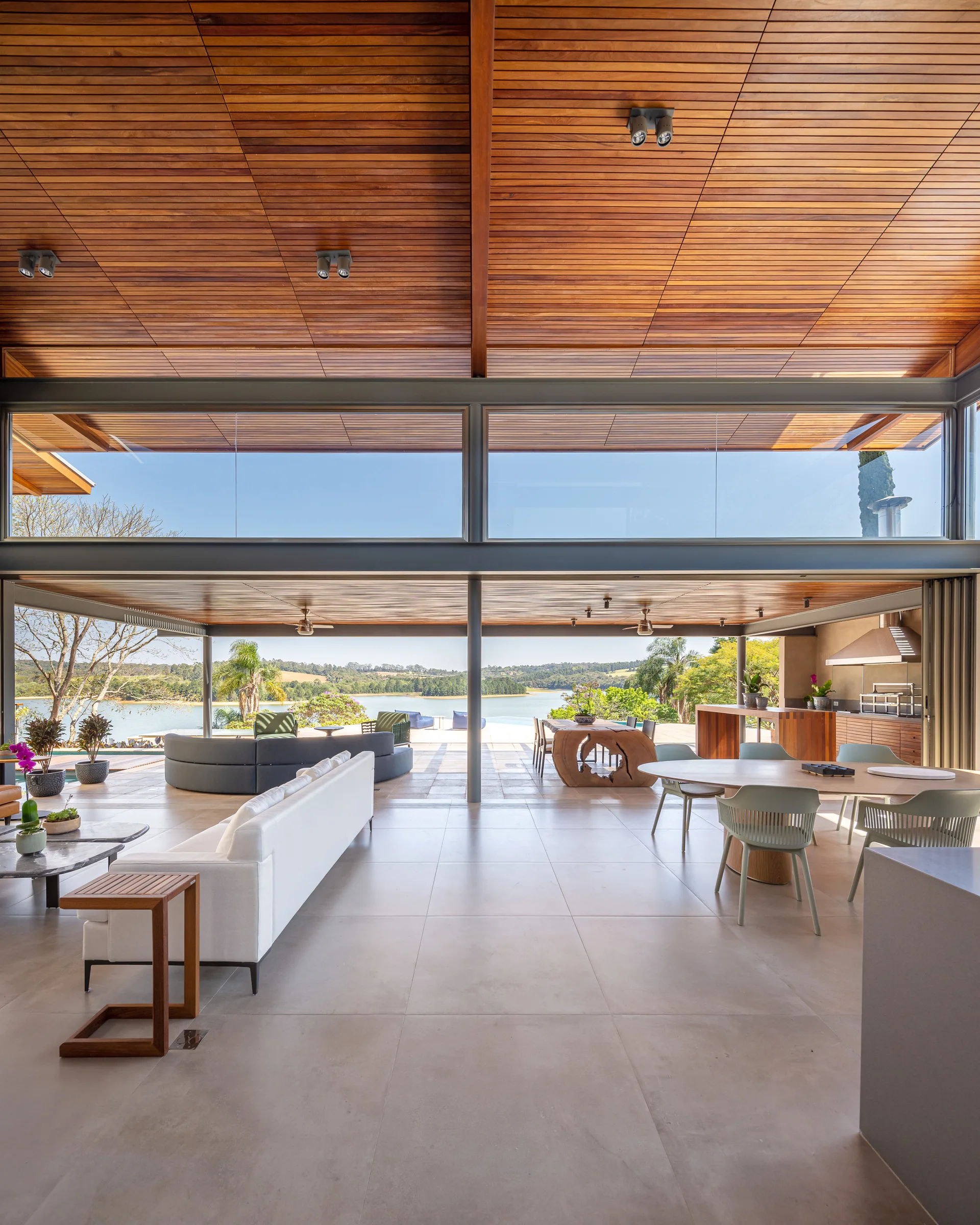 An open-concept living area features a slatted wood ceiling, a white sectional sofa, a large dining table, and massive glass walls overlooking a lake.