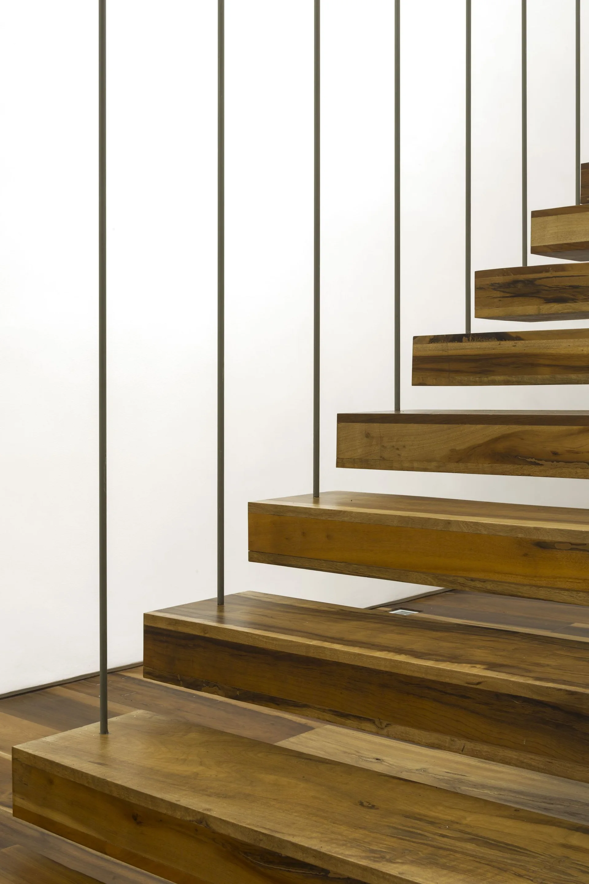 A close-up of a modern floating staircase featuring thick wooden treads and thin vertical metal balusters against a plain white wall.
