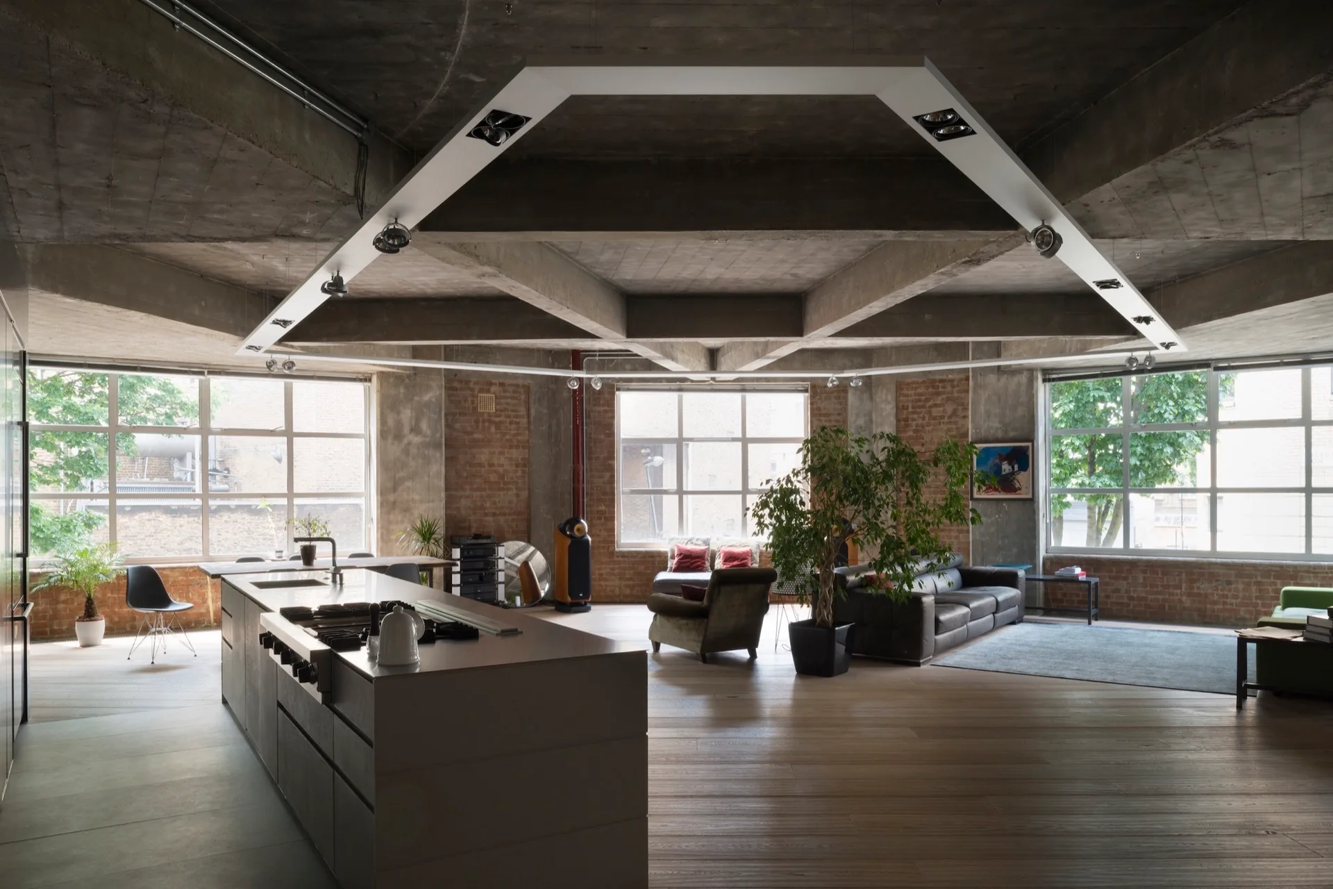An open-concept industrial loft features raw concrete ceilings, exposed brick walls, a minimalist kitchen island, and large grid windows.