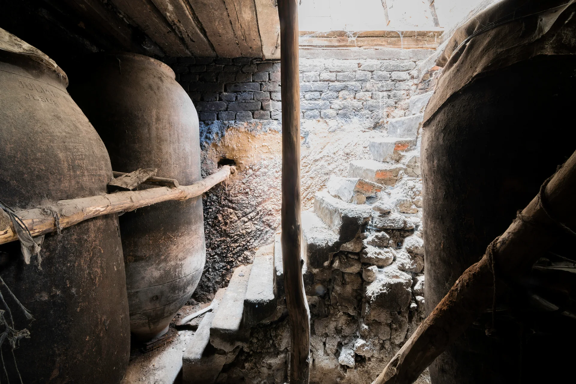 A rustic stone cellar features large clay storage vessels flanking a rough-hewn stone staircase illuminated by natural light from above.