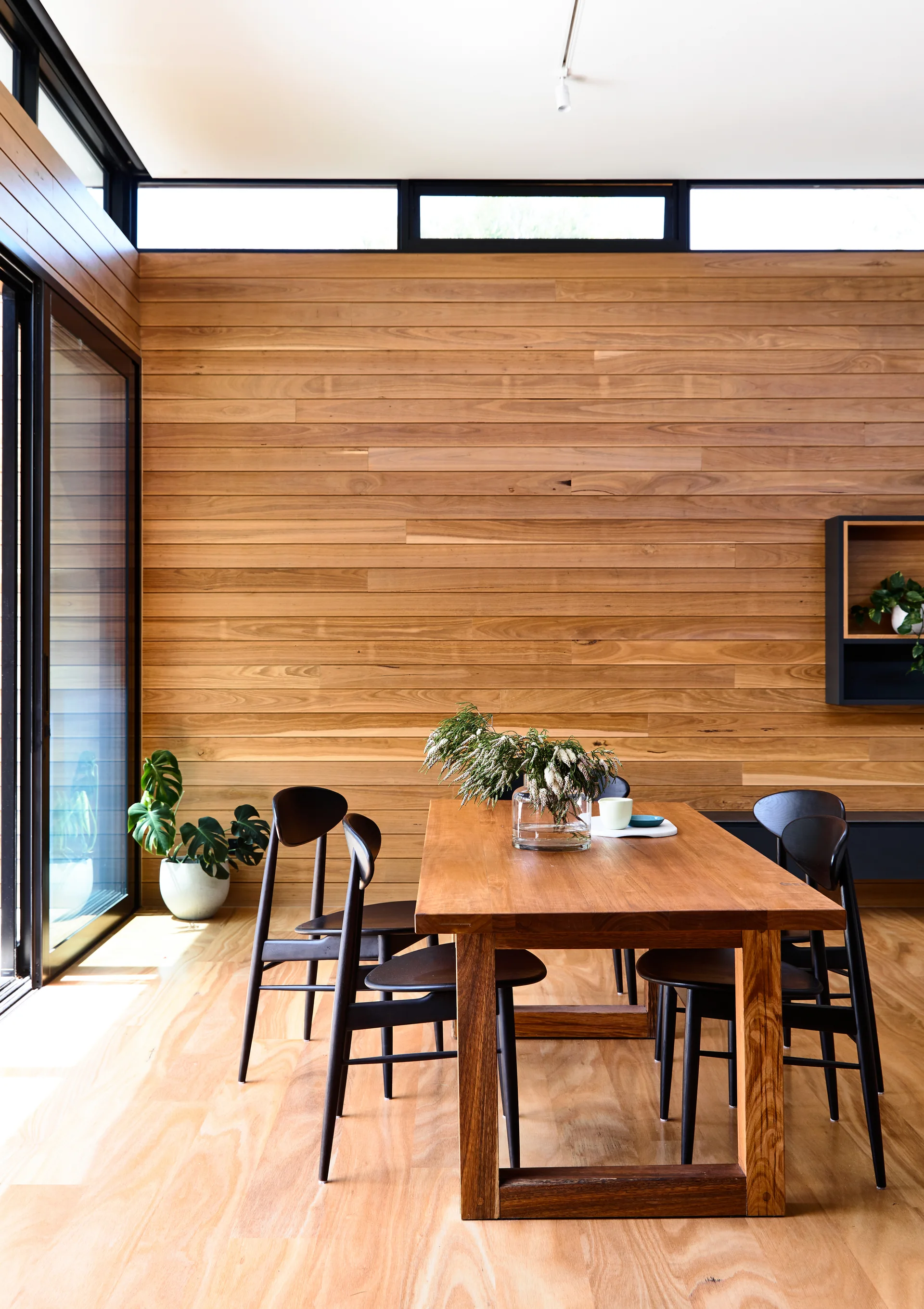 A wooden dining table and matte black chairs sit in a bright room with horizontal wood paneling, large glass doors, and clerestory windows.
