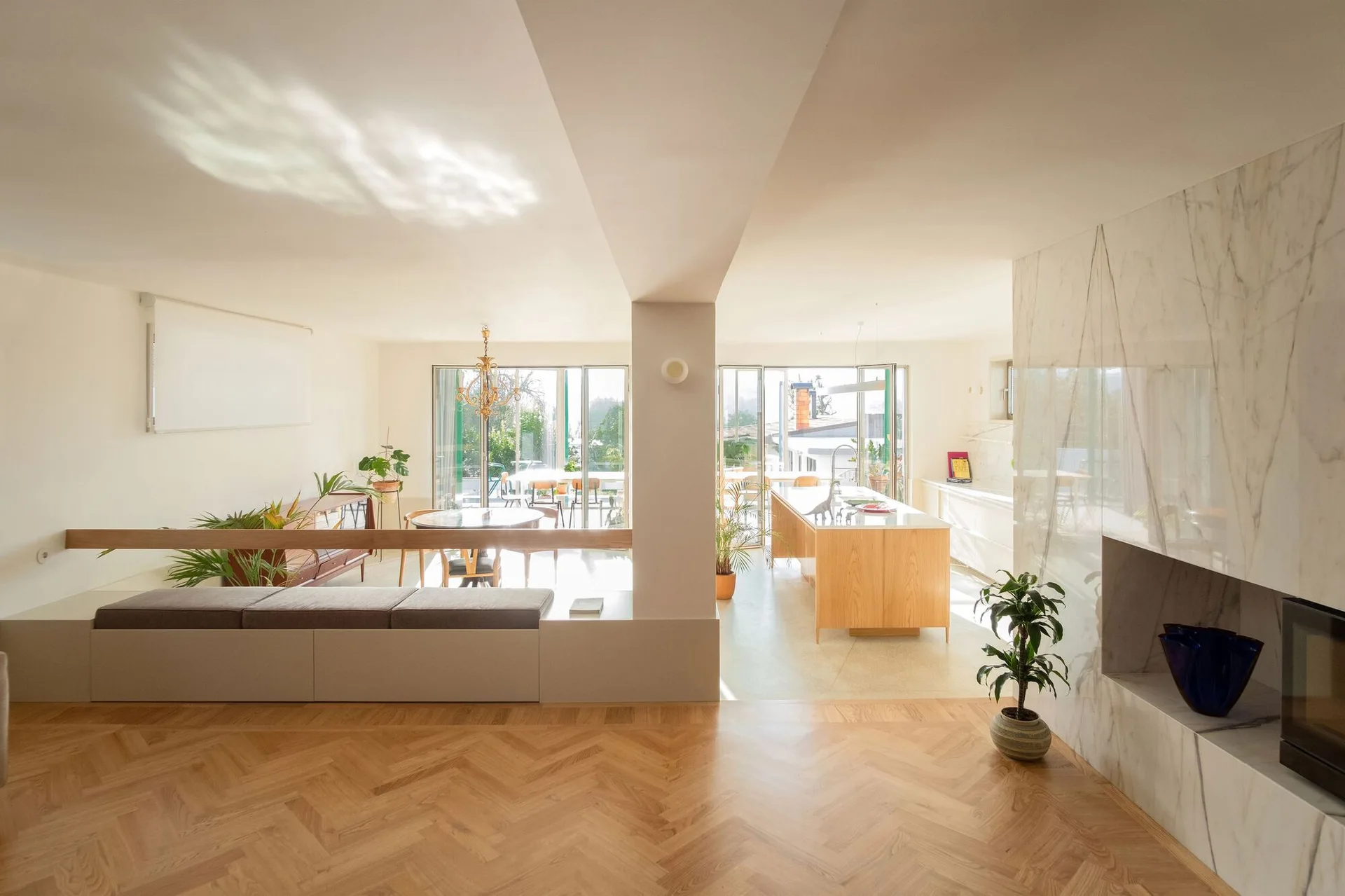 A bright, open-concept living space featuring light herringbone wood floors, a marble fireplace surround, and a low built-in bench.
