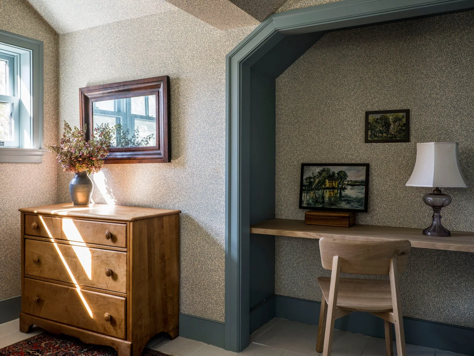 A wooden dresser and a built-in desk nook set against leaf-patterned wallpaper with blue-gray painted trim and a matching wooden chair.