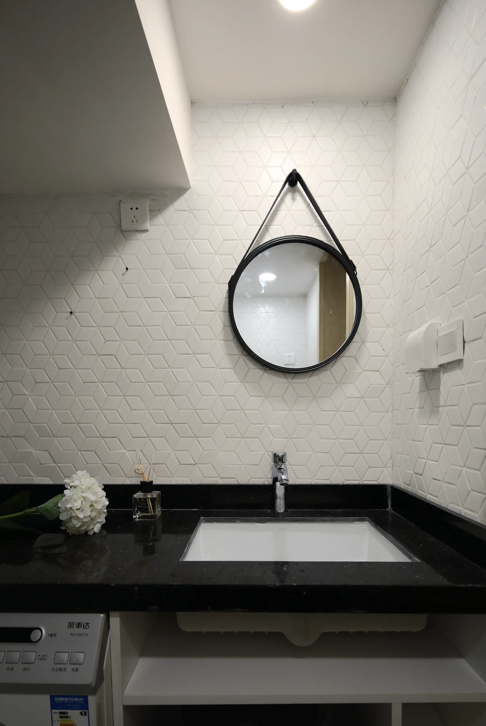 A bathroom vanity with a black countertop and sink below a round black-framed mirror hanging against a white geometric tiled wall.