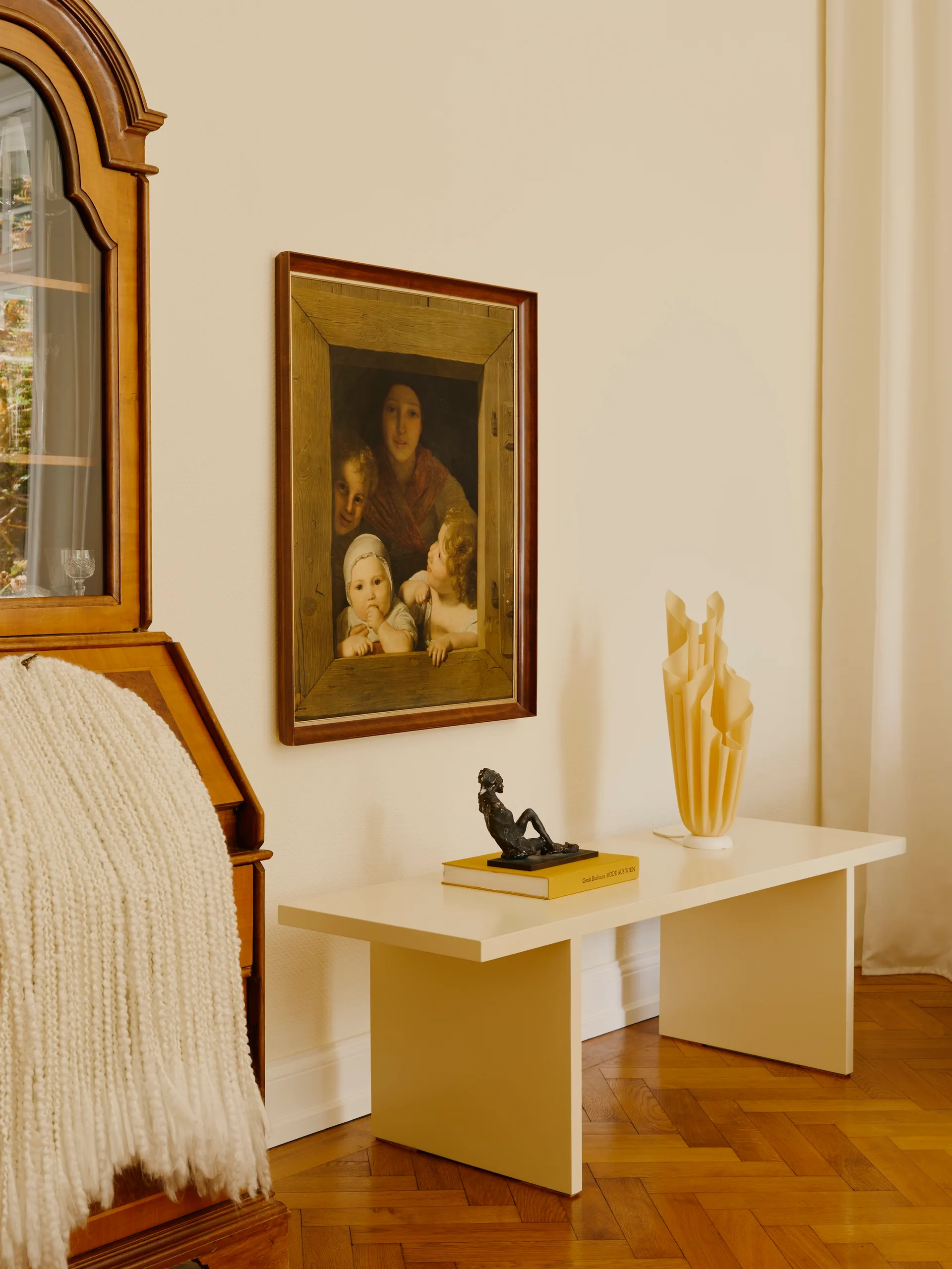 A minimalist cream console table holding a sculptural lamp and small sculpture stands next to a vintage wooden secretary desk.