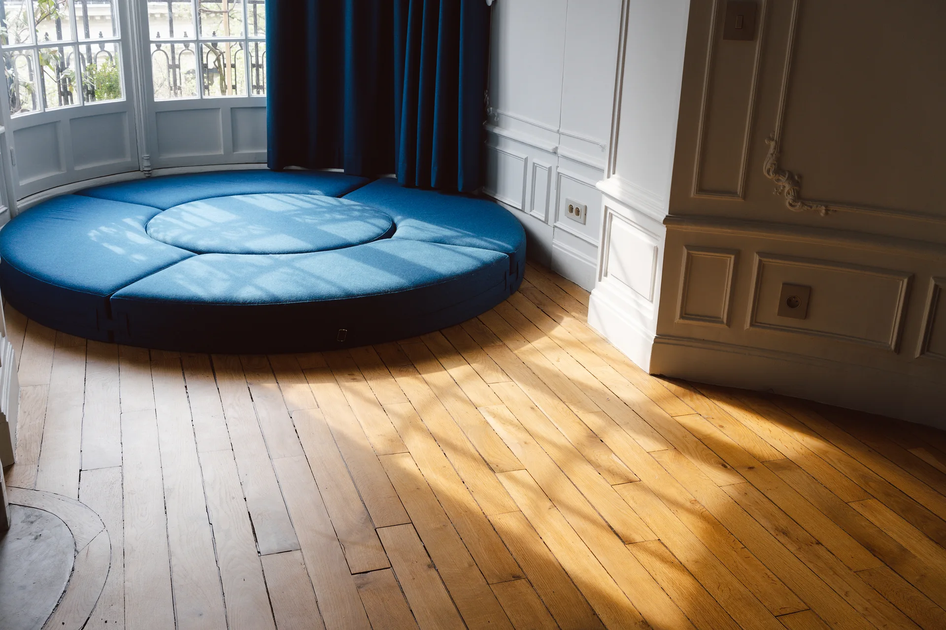 A circular blue fabric floor bed sits near a bay window with matching blue curtains and white wainscoting on a light hardwood floor.