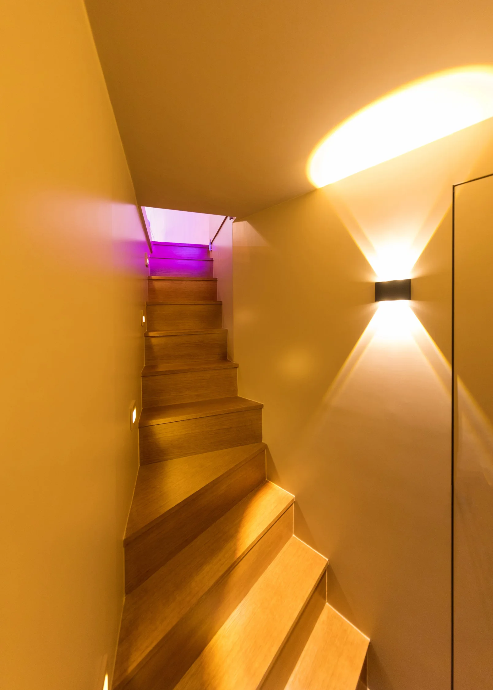 A narrow wooden staircase with beige walls features step lights on one side and a black up-and-down wall sconce casting beams of light.
