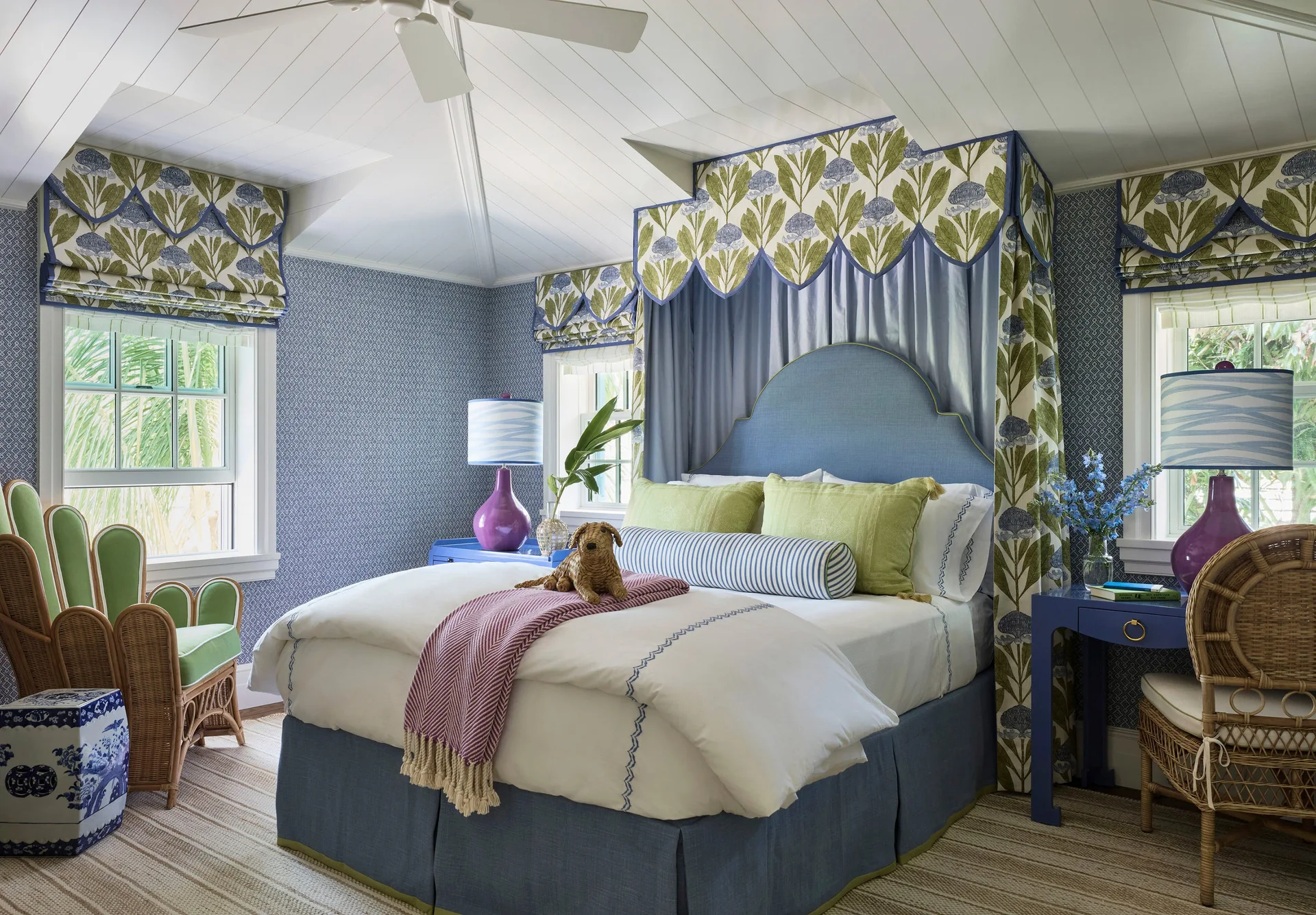 A bedroom features a blue canopy bed with a botanical fabric valance, small-scale patterned wallpaper, and wicker chairs.