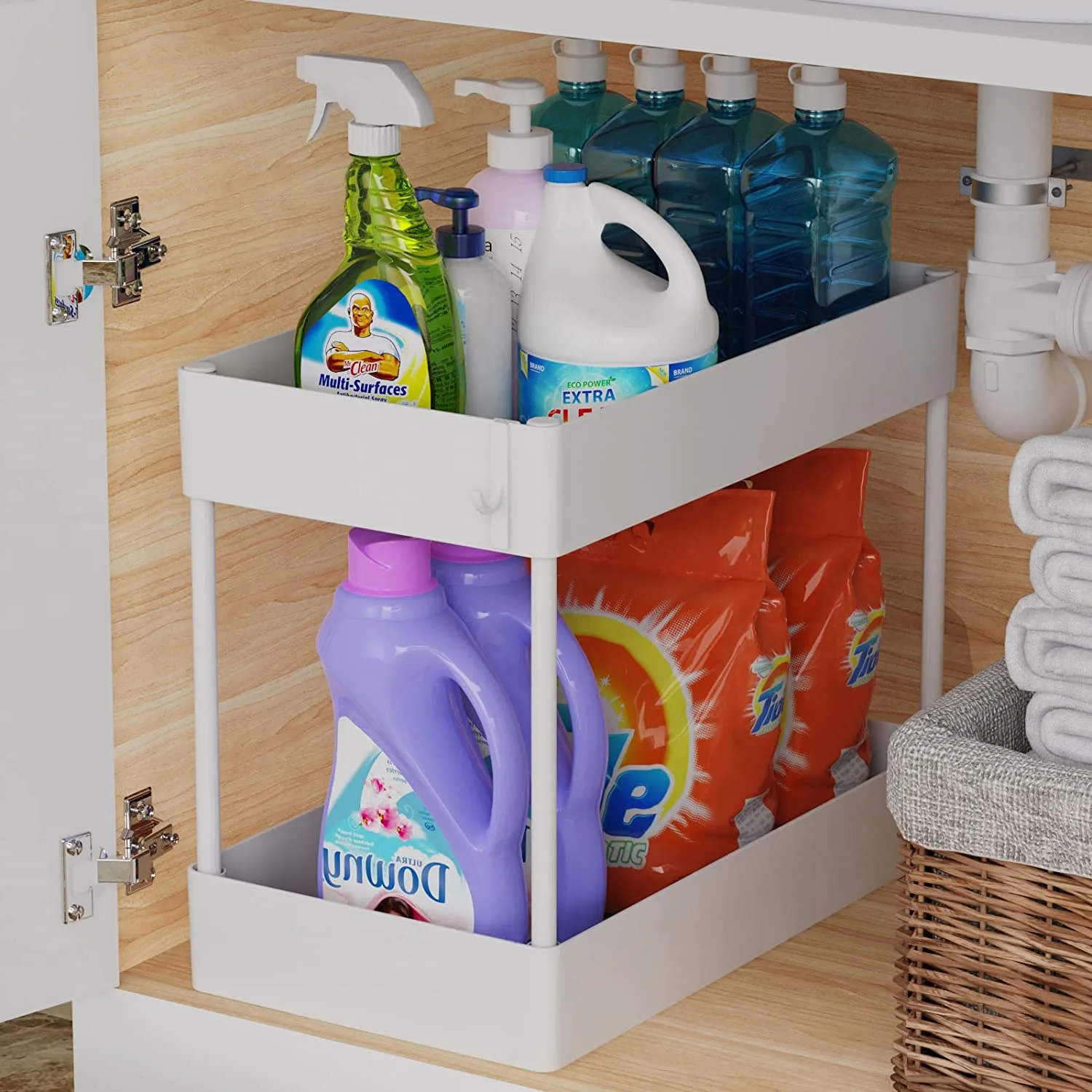 A two-tier white plastic organizer filled with cleaning supply bottles sits inside a light wood-grained cabinet next to plumbing pipes.