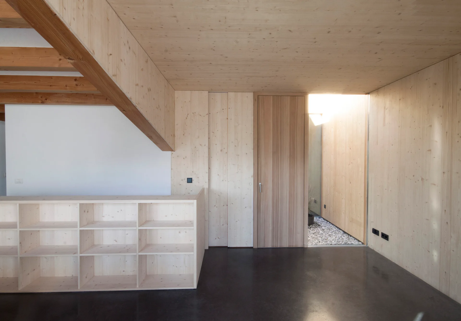 A minimalist interior space features walls and ceilings clad in light wood, a low wooden shelving unit, and a dark polished concrete floor.