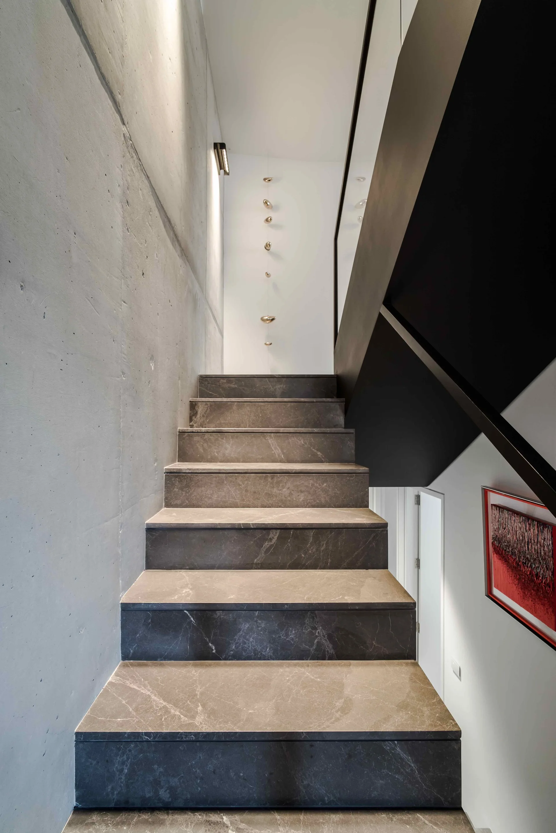 A modern staircase featuring grey veined stone steps is bordered by a raw concrete wall and a minimalist black metal handrail.