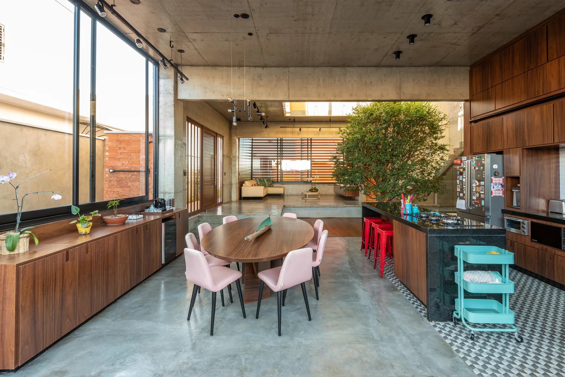 An open-plan industrial kitchen features an oval wood table with pink chairs, wood cabinetry, and exposed concrete ceilings.