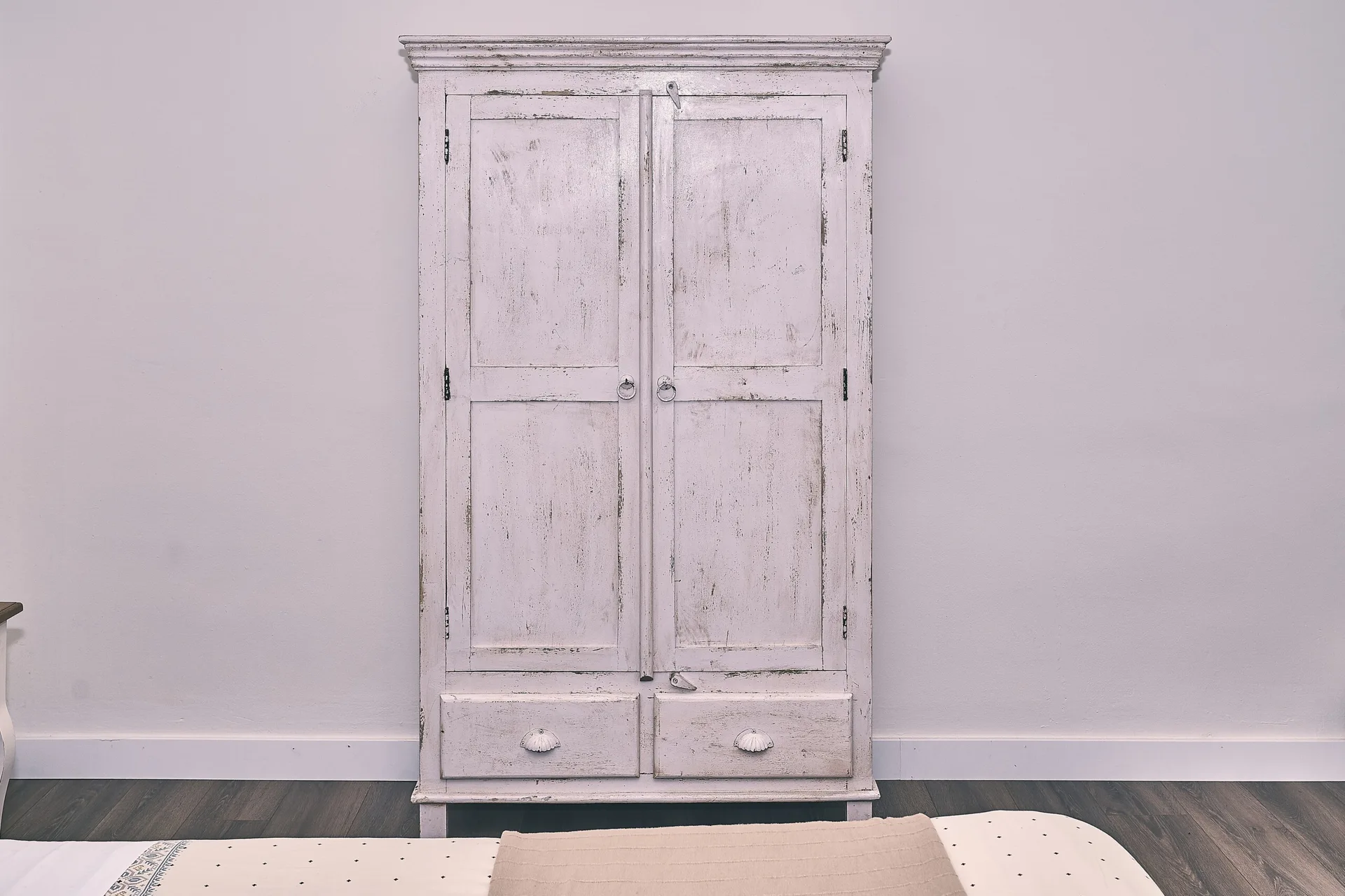 A tall, distressed white wooden wardrobe with double doors and two bottom drawers stands against a plain light wall on dark wood floors.