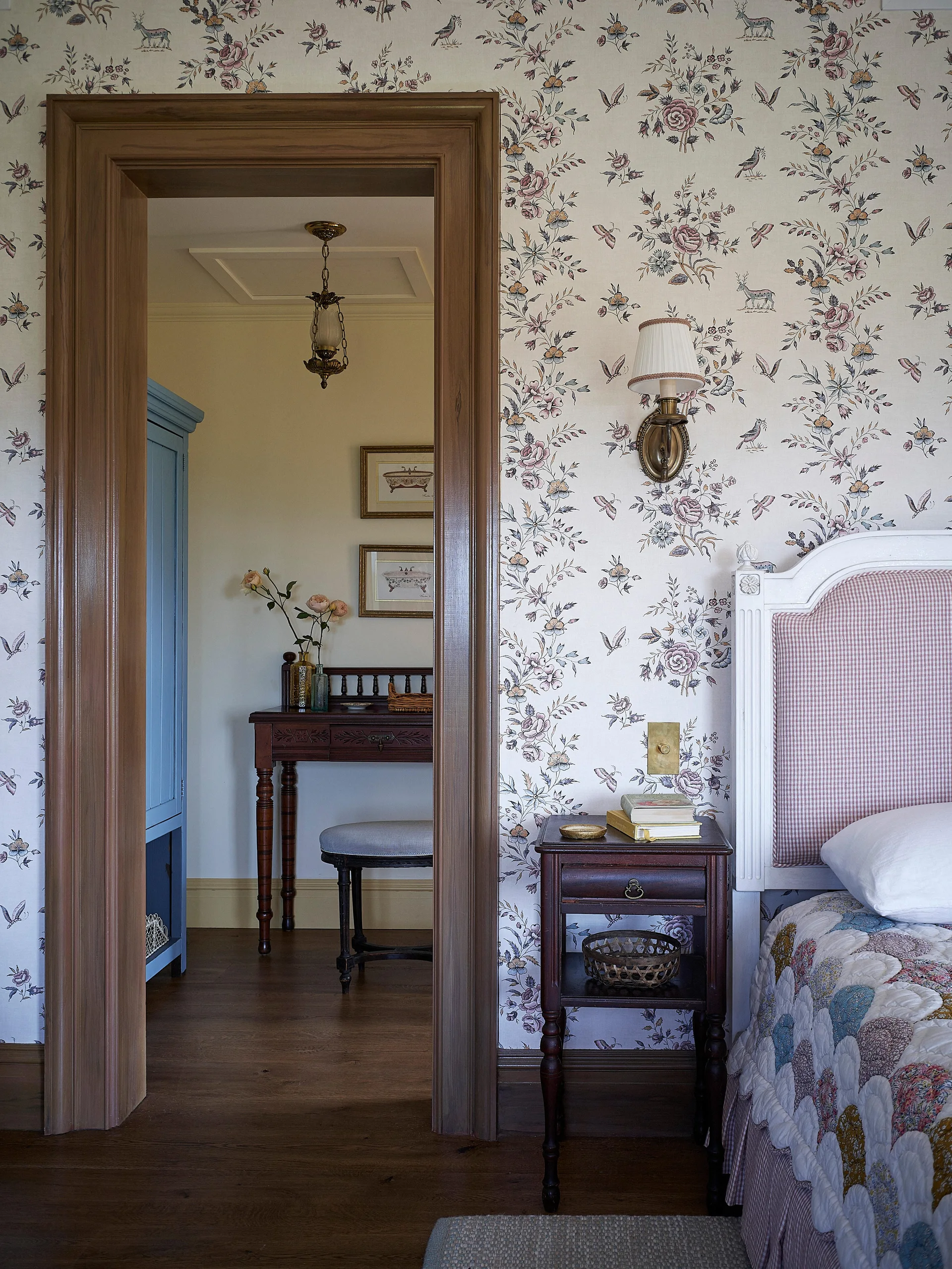 A cottage bedroom features floral pattern wallpaper, a wooden doorway, a dark wood nightstand, and a glimpse into an adjacent dressing room.