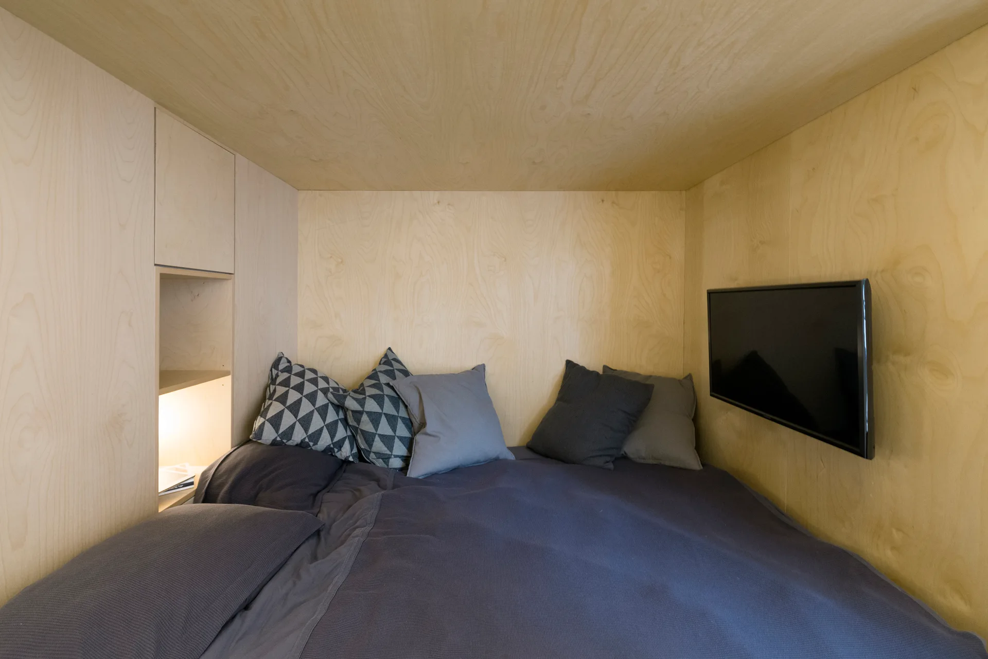 An enclosed sleeping nook lined in light wood veneer panels with a grey-dressed bed, a wall-mounted television, and a built-in lighted shelf.