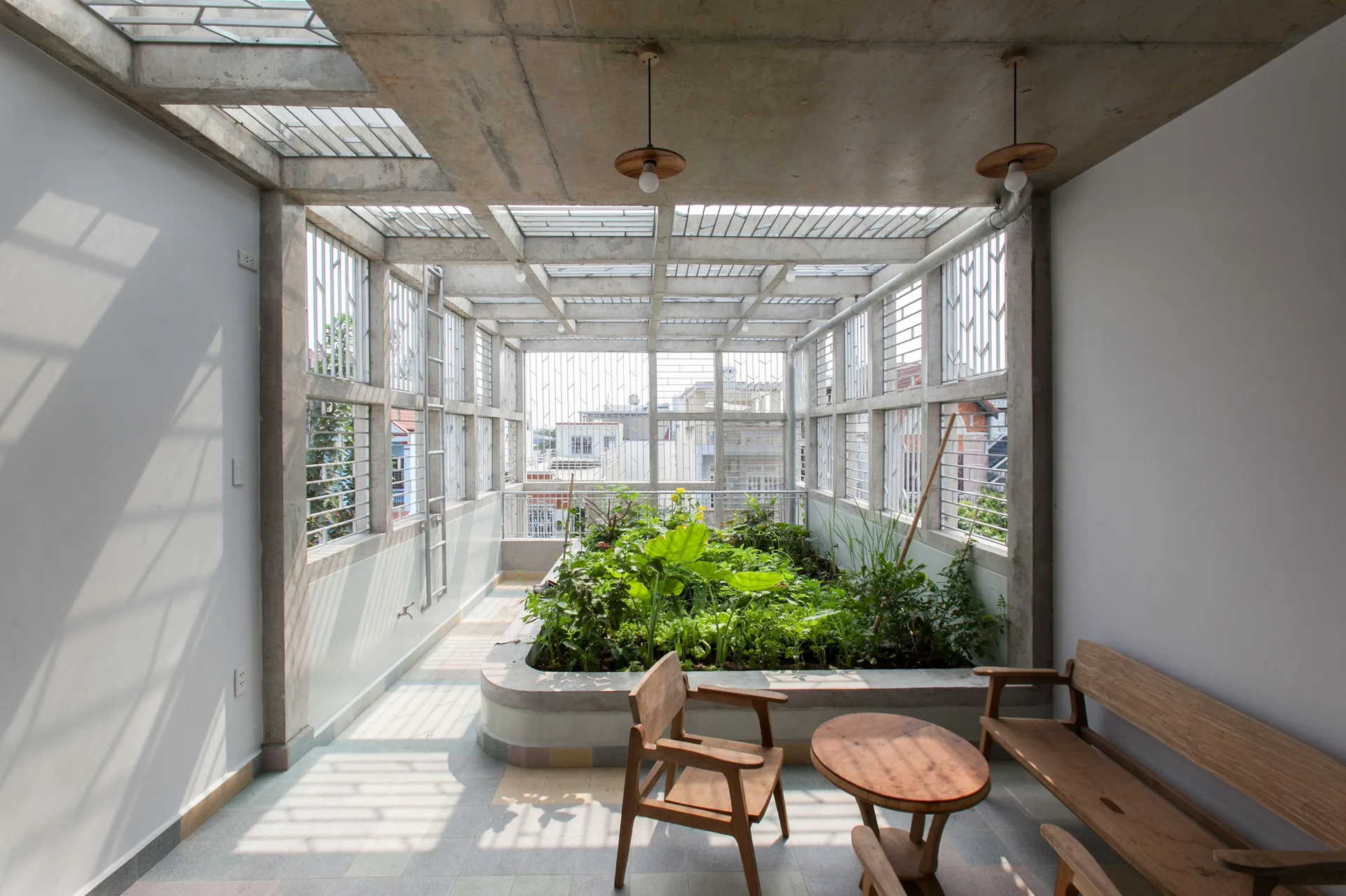 A semi-outdoor concrete terrace features a central built-in garden planter, wooden furniture, and a gridded metal skylight roof.