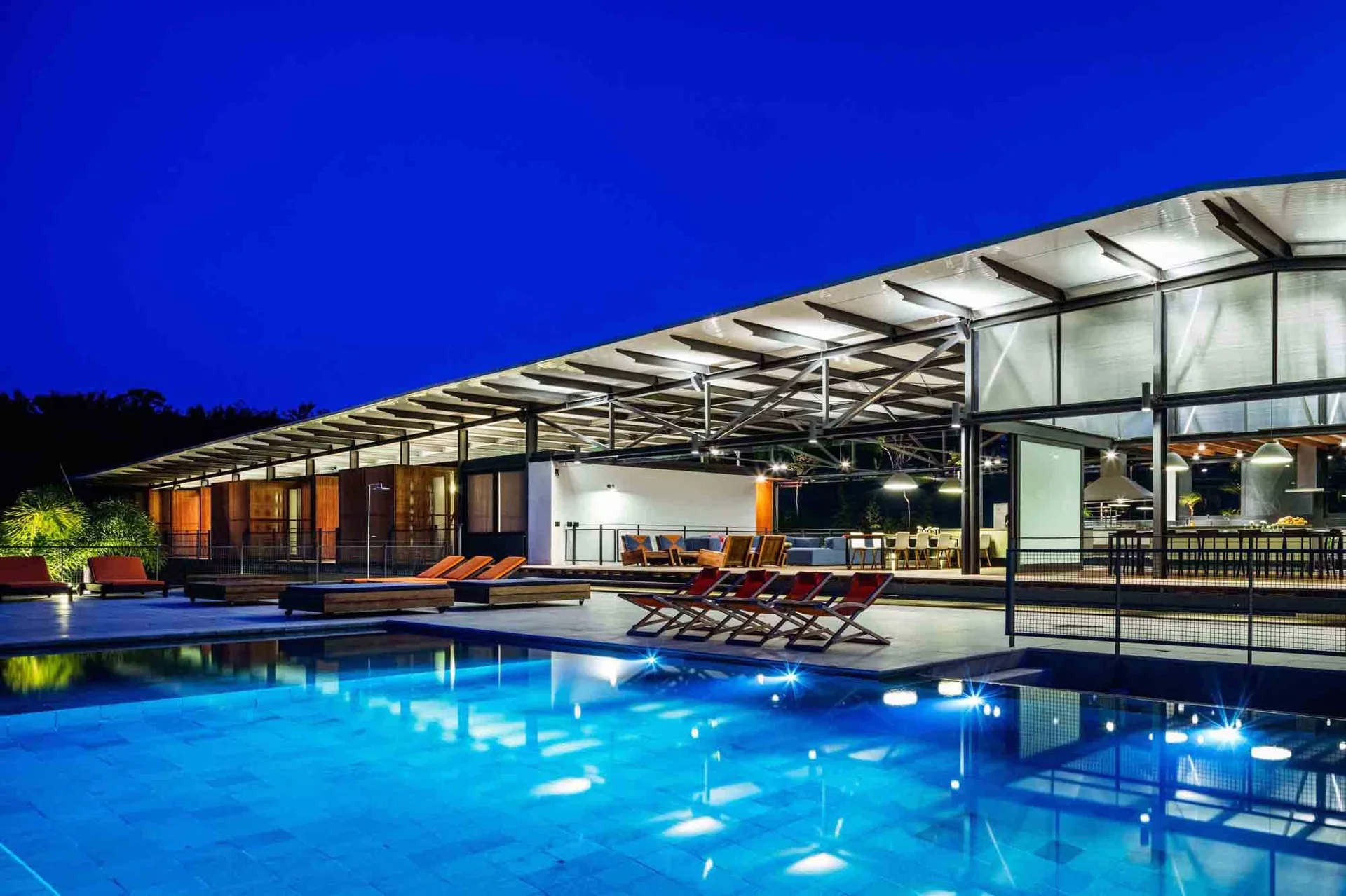 A large illuminated swimming pool sits next to a modern covered patio with an exposed steel roof, dining furniture, and orange lounge chairs.