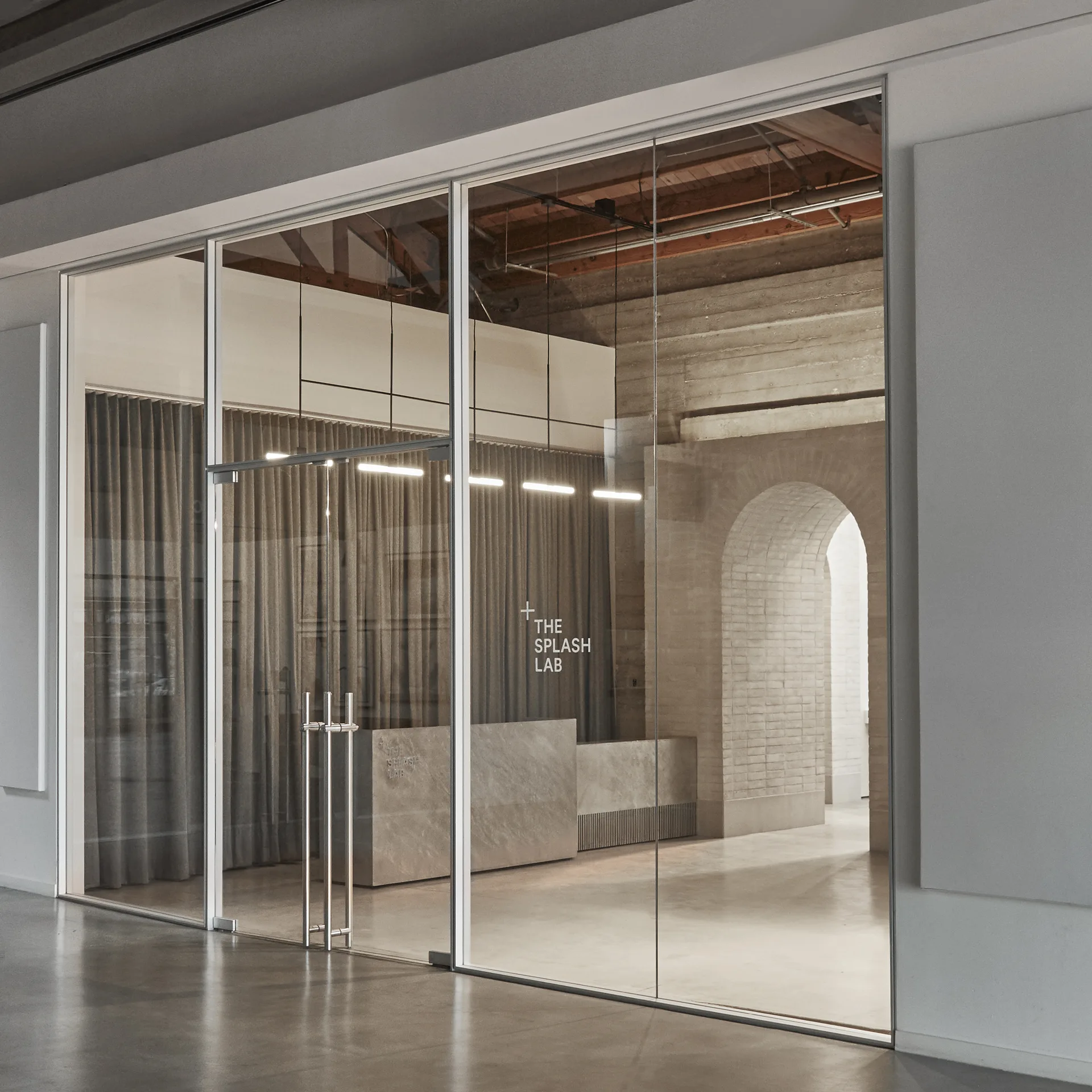 An industrial reception entryway viewed through a glass wall, featuring a stone desk, a fabric curtain backdrop, and a brick archway.
