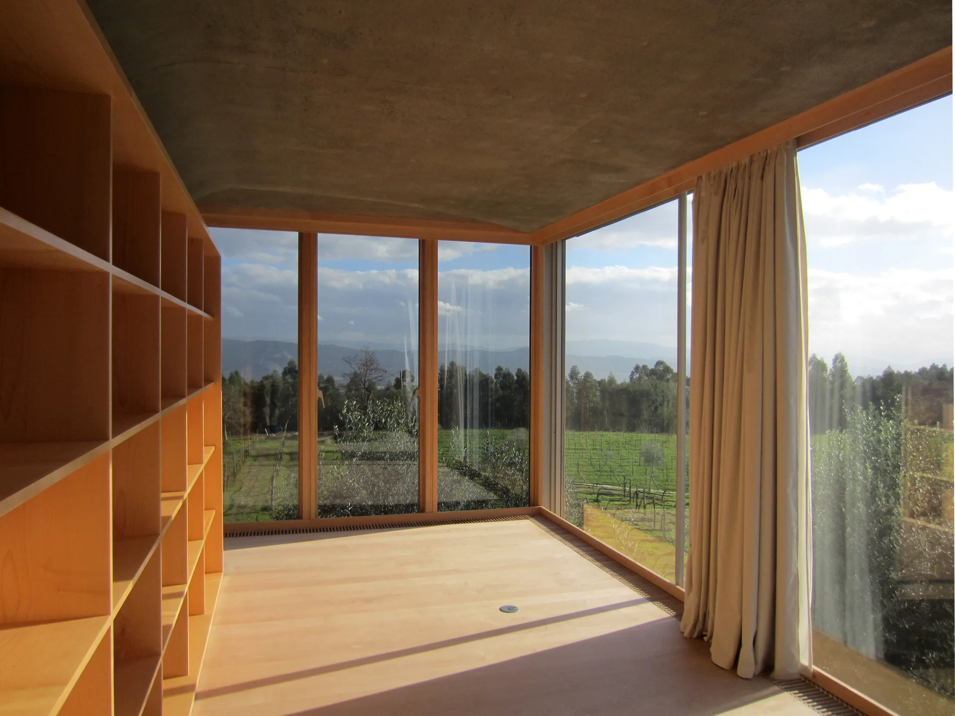 A minimalist room featuring a large light wood bookshelf, exposed concrete ceiling, and floor-to-ceiling windows showing a green landscape.