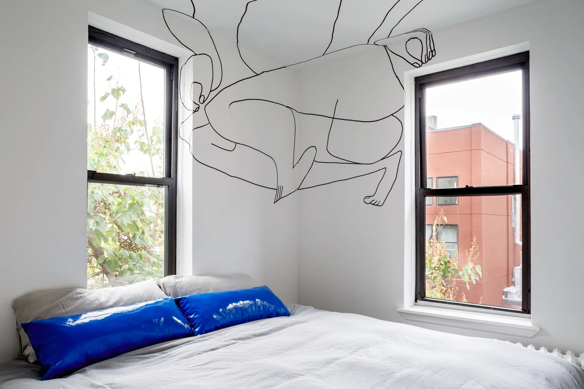 A bedroom corner features a bed with shiny blue bolster pillows and a black line drawing painted on the wall between two windows.
