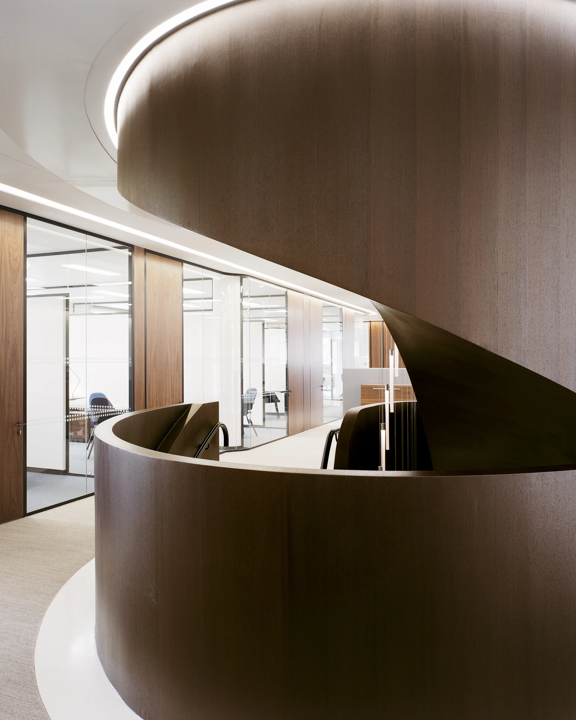 A large curved staircase clad in dark wood stands in an office with glass partitions and recessed curved ceiling cove lighting.