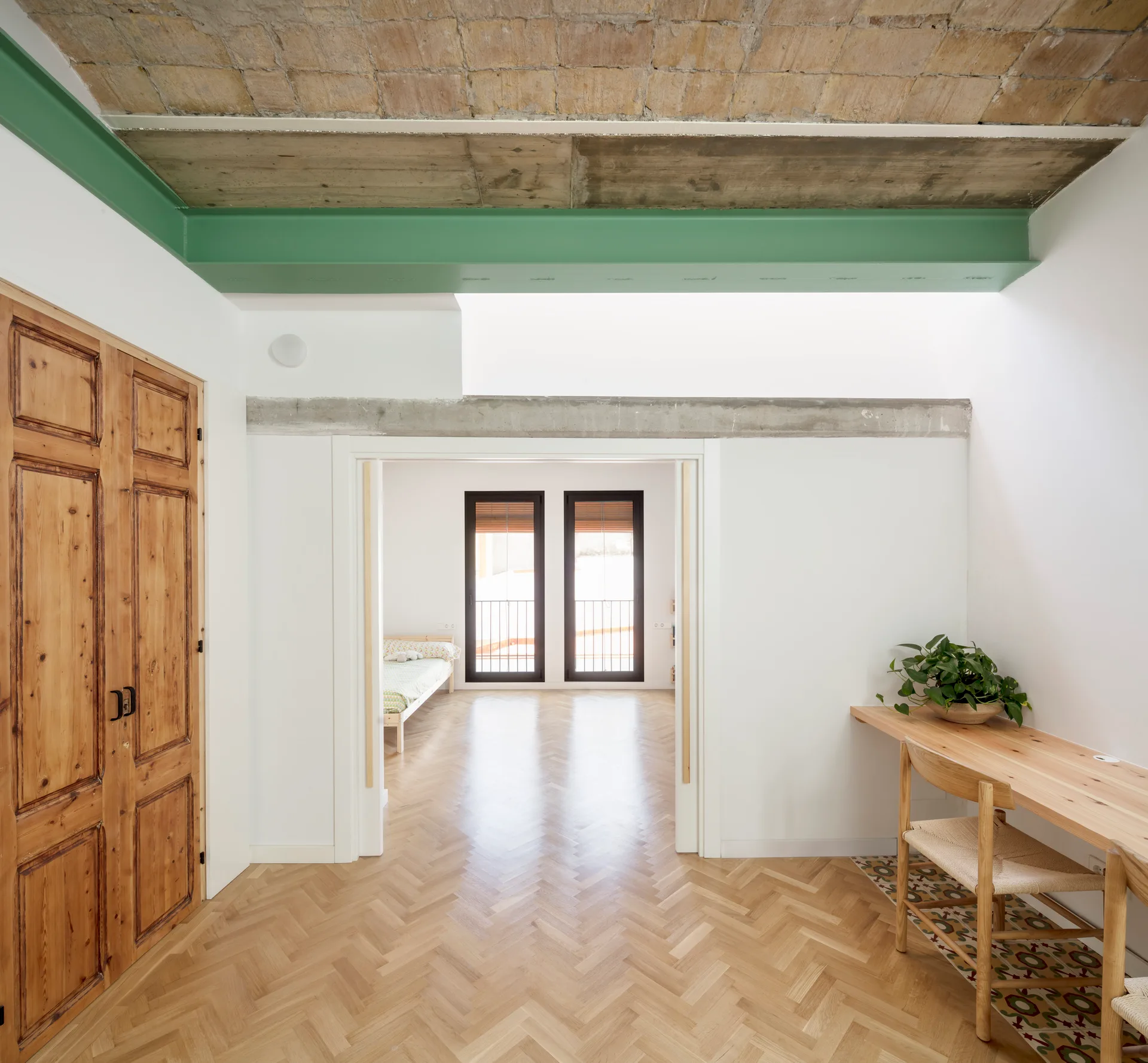 A bright hallway features herringbone wood floors, a green painted ceiling beam, and a wooden desk leading toward a bedroom with double glass doors.