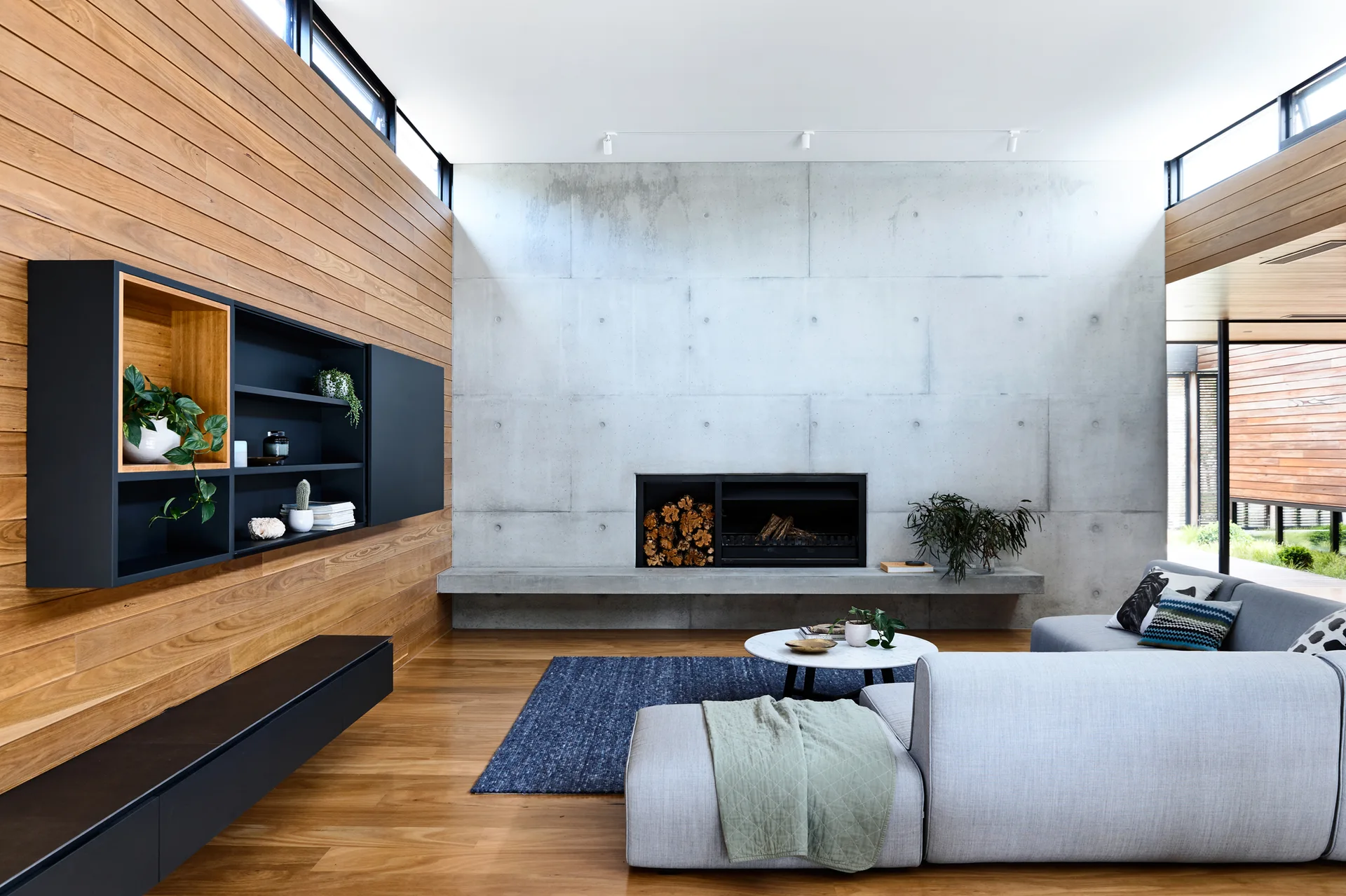 A modern living room featuring a concrete fireplace feature wall, warm horizontal wood paneling, a gray sectional sofa, and a black wall shelving unit.