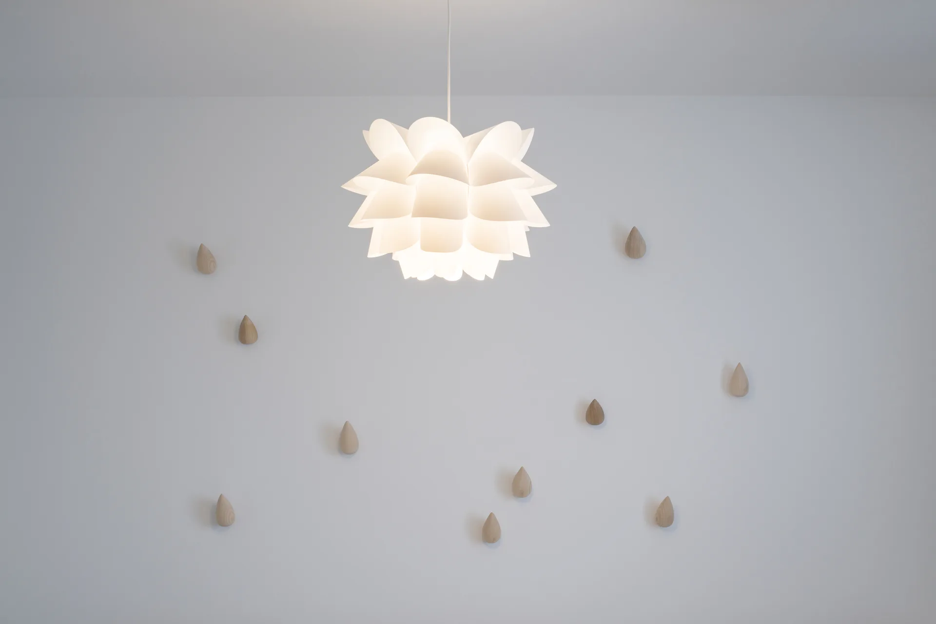 A white sculptural pendant light hangs in front of a white wall decorated with scattered wooden drop-shaped pegs.