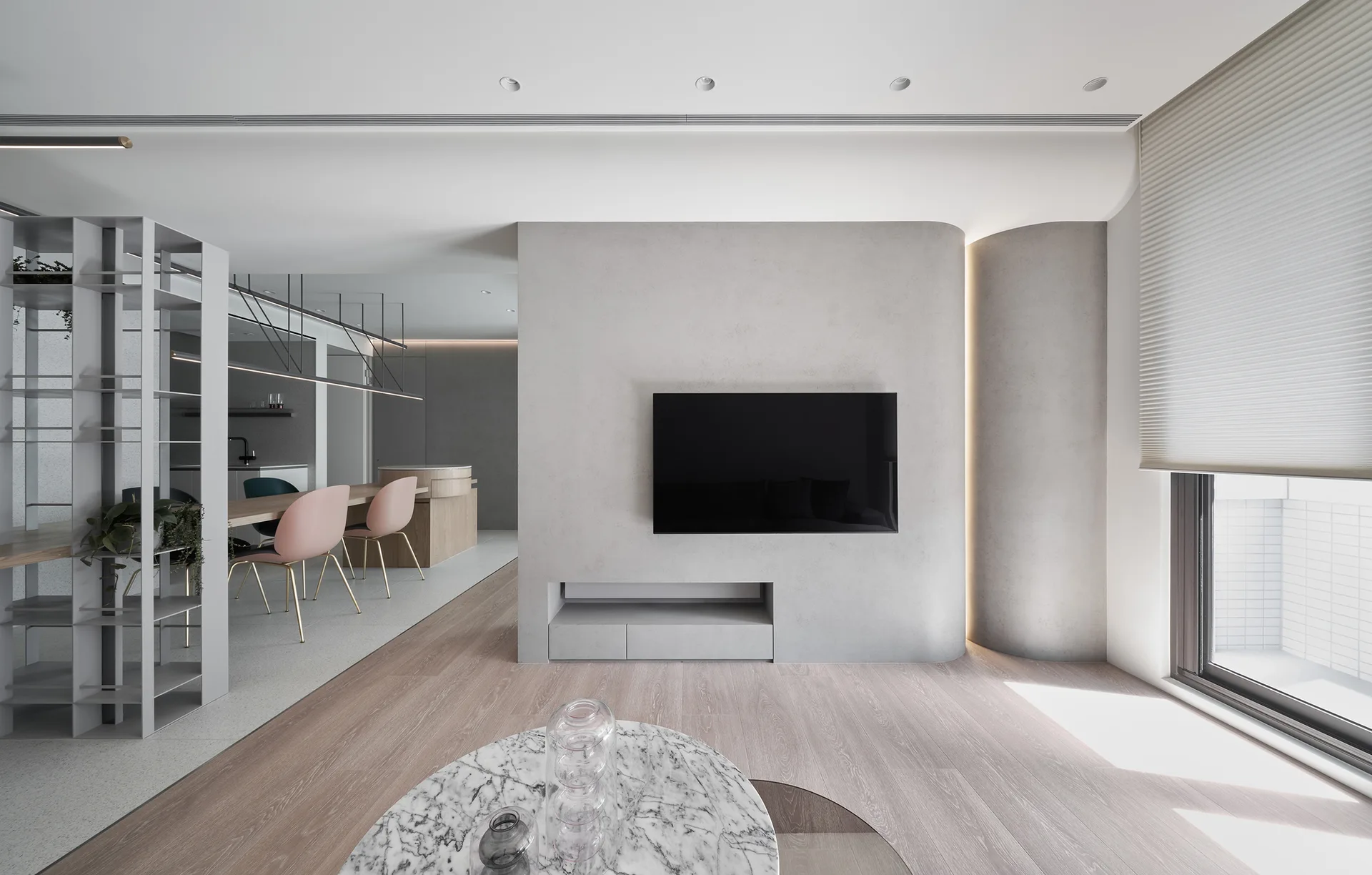 A minimalist living room features a curved gray plaster feature wall with a television, light wood floors, and a dining table with pink chairs behind it.