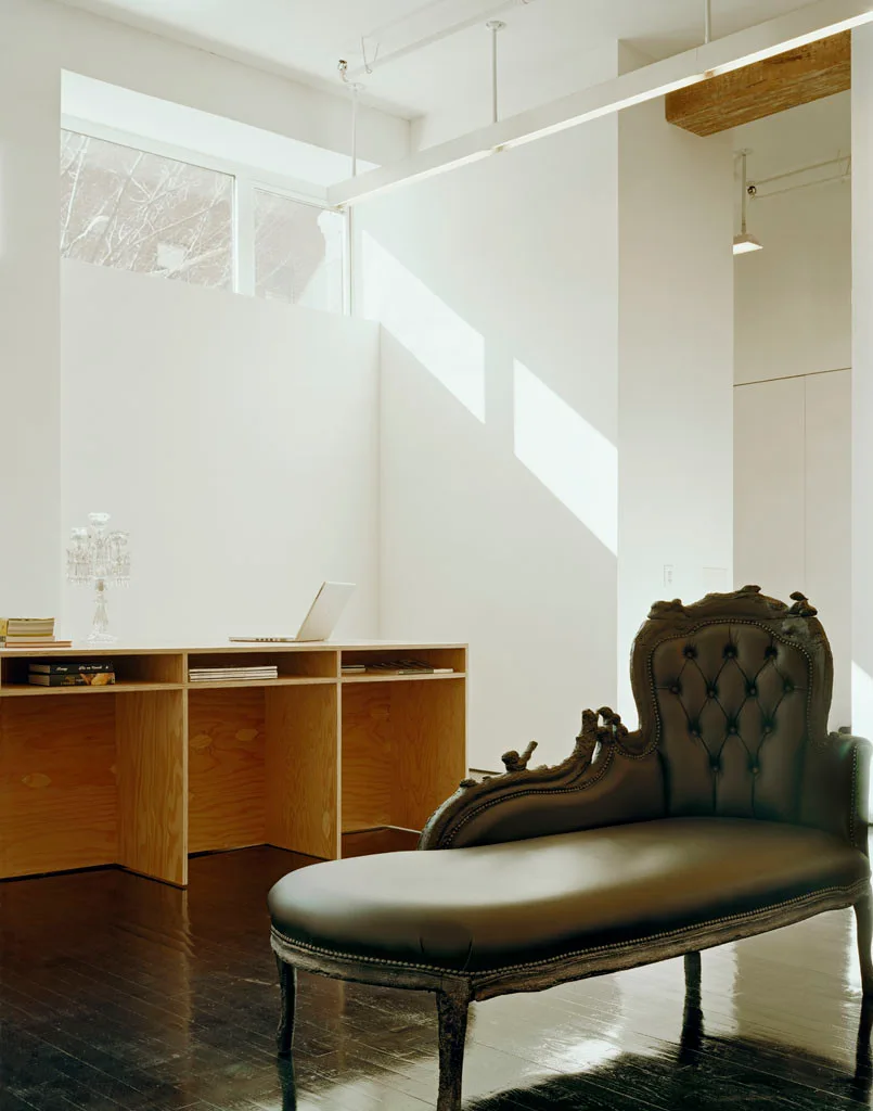 A minimalist home office featuring a plywood desk and an ornate black tufted leather chaise longue on dark glossy wood floors.