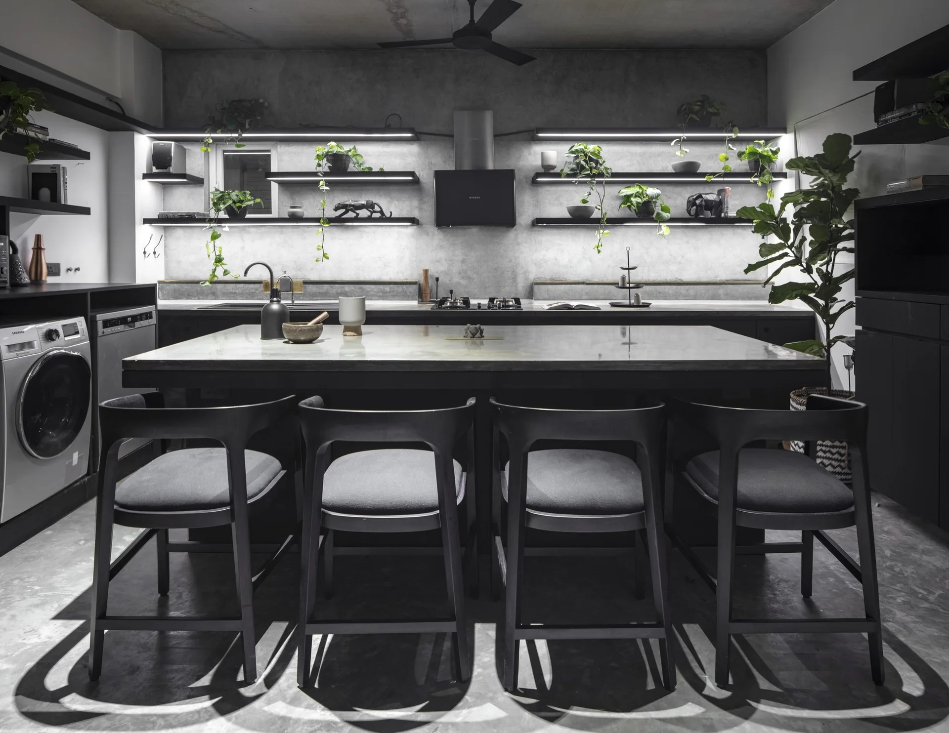A modern industrial kitchen featuring a concrete island with four black bar stools and illuminated floating shelves decorated with green plants.