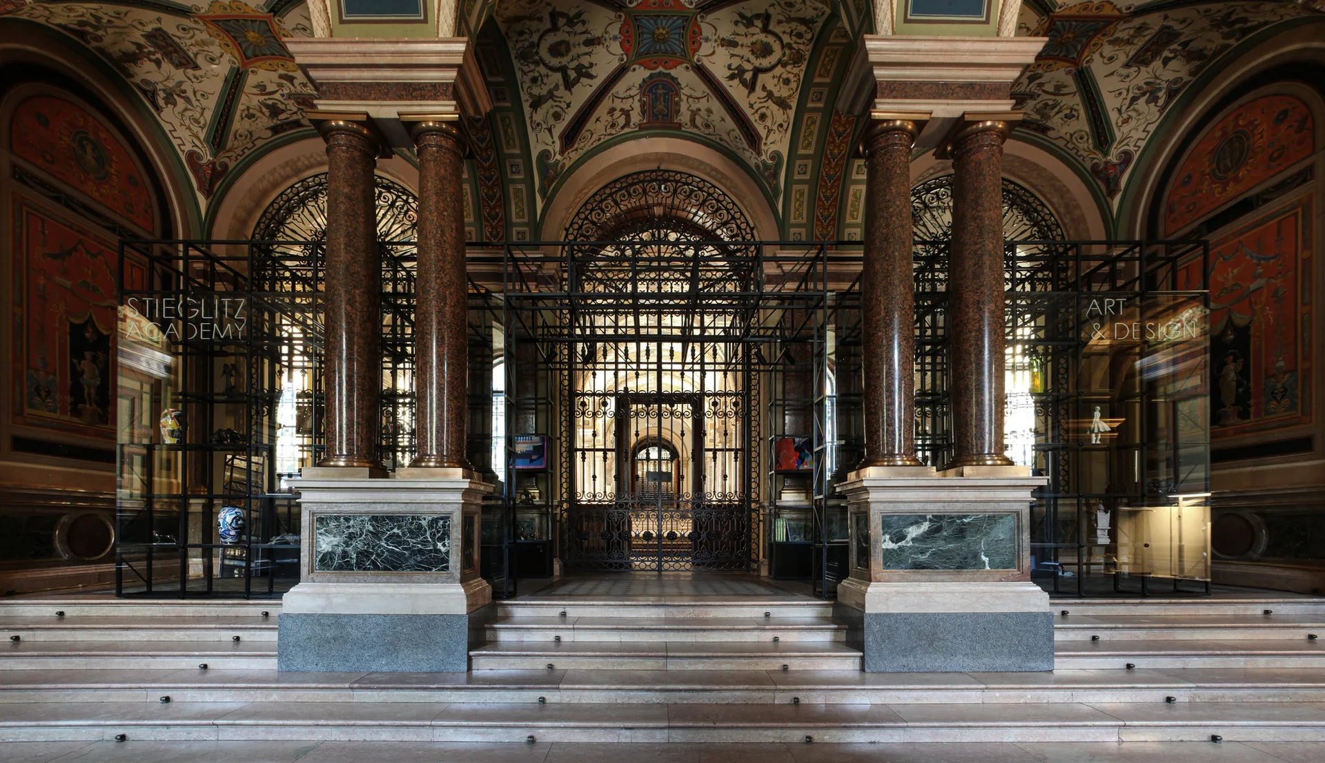 A grand entrance hall features massive polished stone columns on marble bases, an ornate painted vaulted ceiling, and a central wrought iron gate.