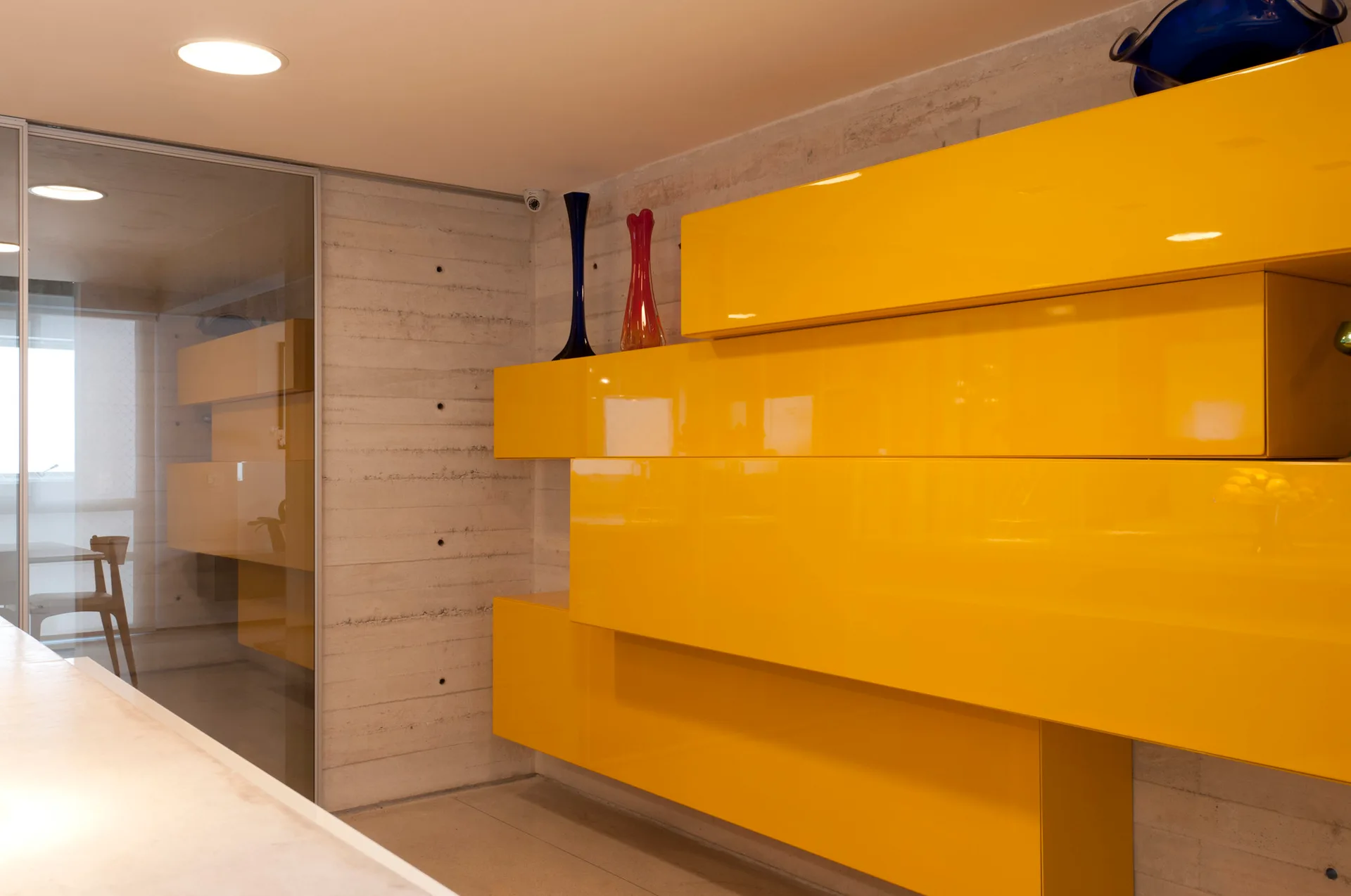 Bright yellow high-gloss modular cabinets are mounted on a raw grey board-formed concrete wall next to a glass partition.