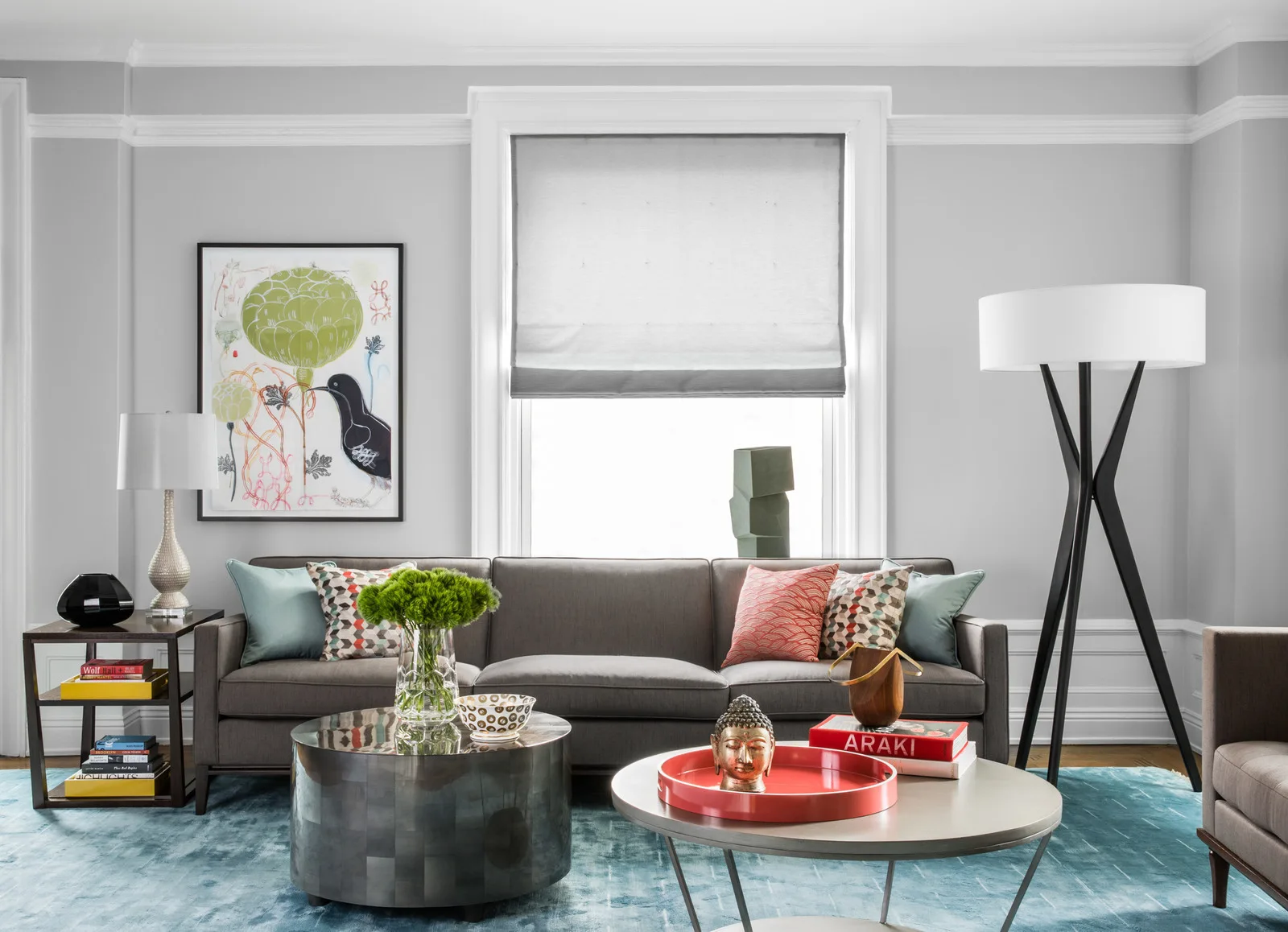 A transitional living room features a grey sofa, a blue patterned area rug, two round coffee tables, and a large window with a grey Roman shade.
