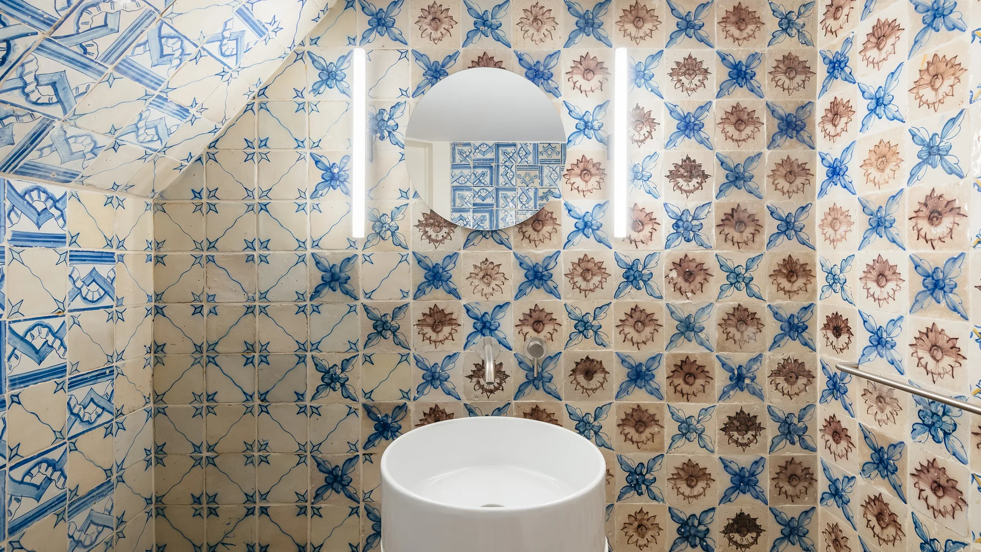 A bathroom features a round mirror and white pedestal sink against walls fully covered in blue and brown patterned ceramic tiles.